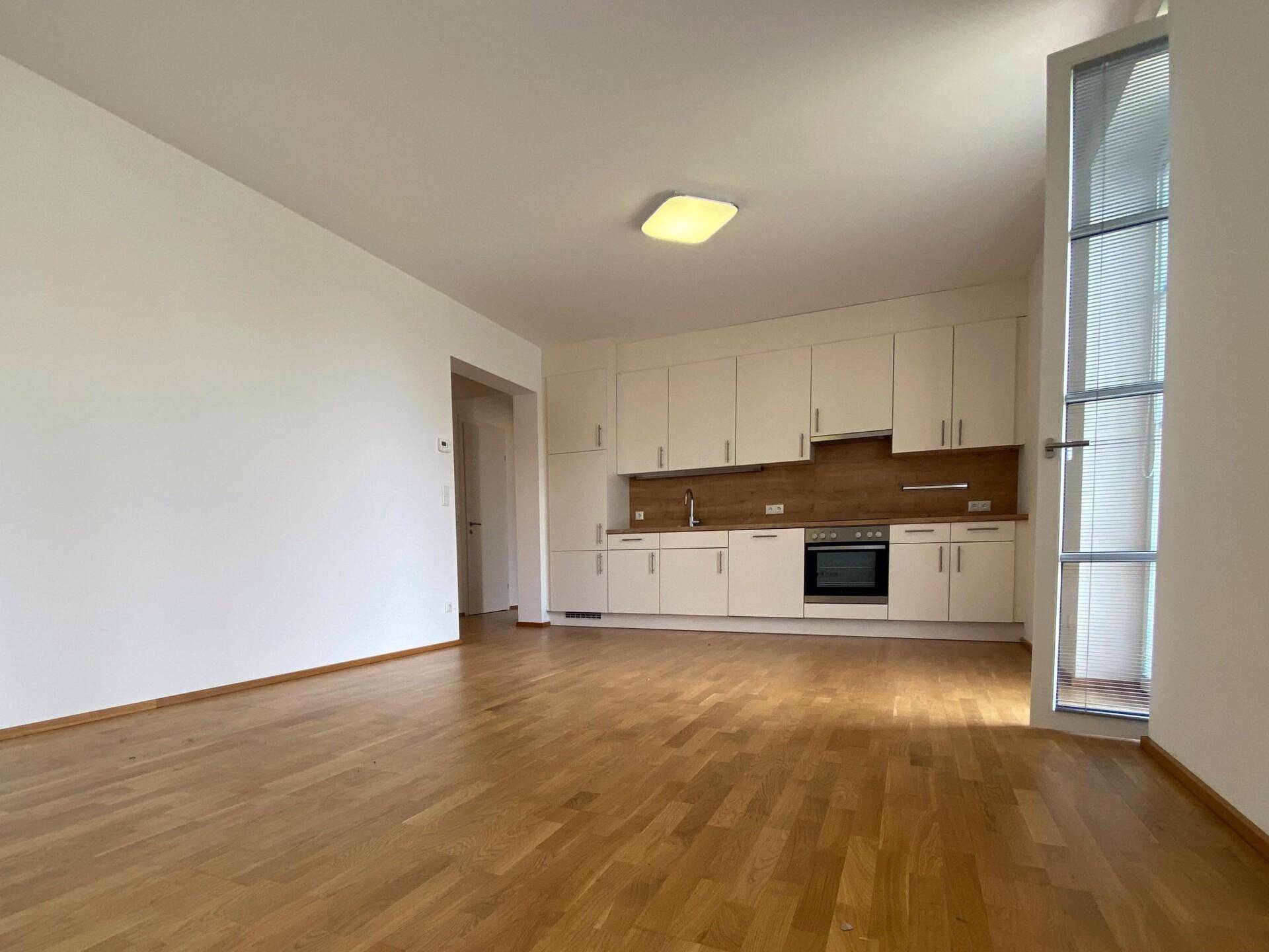 Heller offener Wohnbereich mit integrierter Küchenzeile, Parkettboden und hohem Fenster.