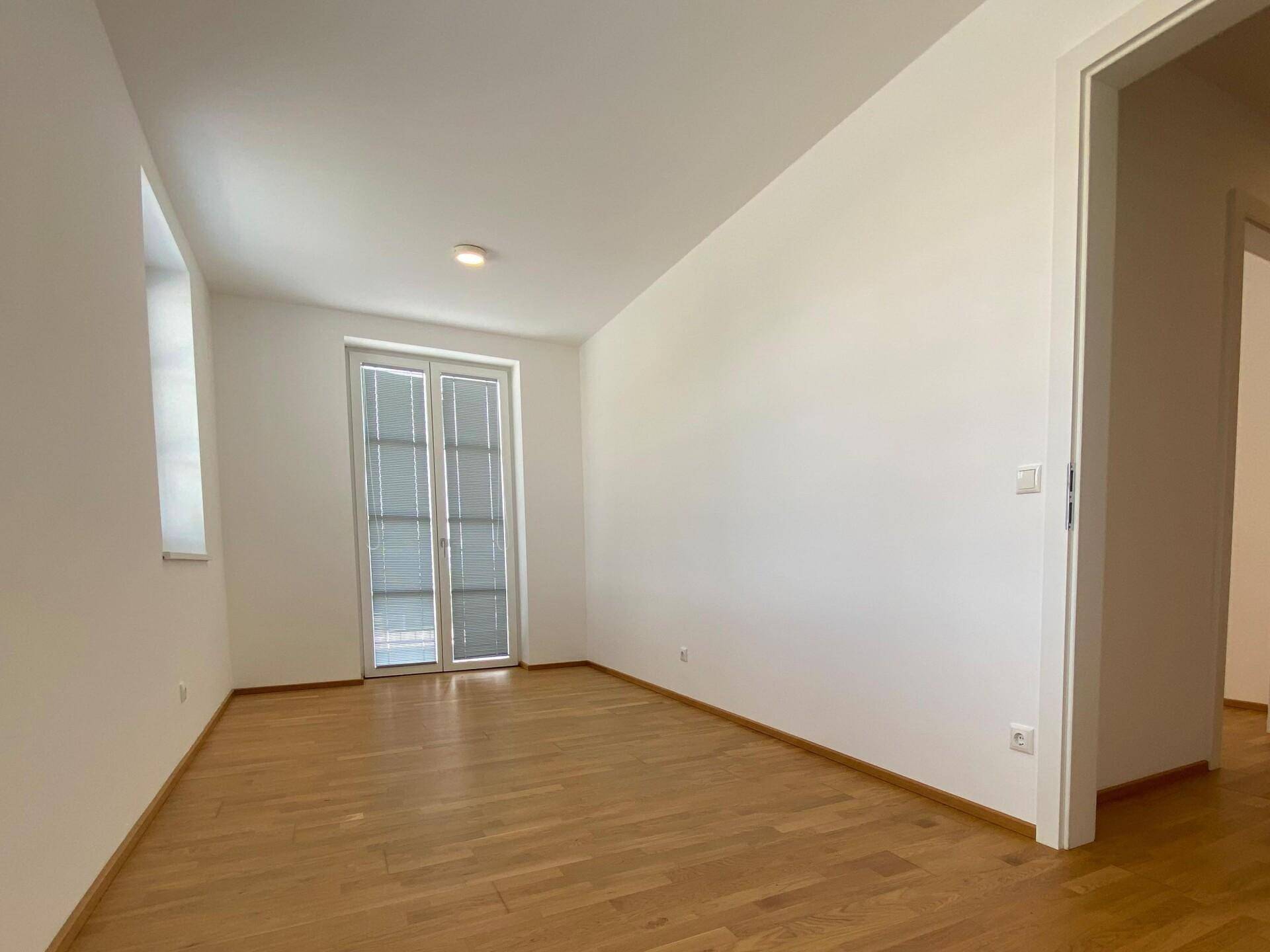 Leerer Raum mit Parkettboden und Doppeltür, die zu einem französischen Balkon oder einer kleinen Terrasse führt.