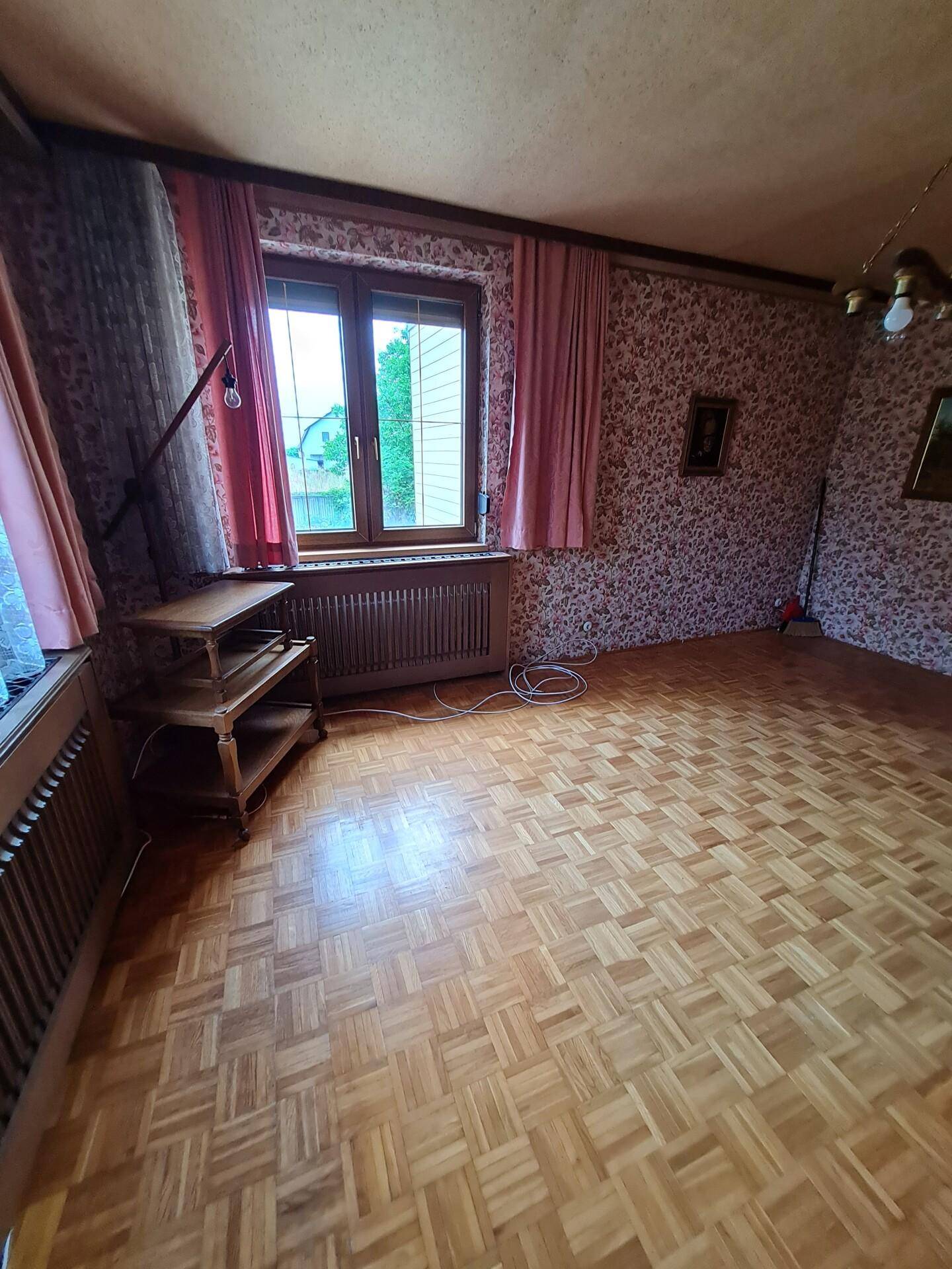 Geräumiges Wohnzimmer mit Parkettboden, floraler Tapete und großem Fenster.