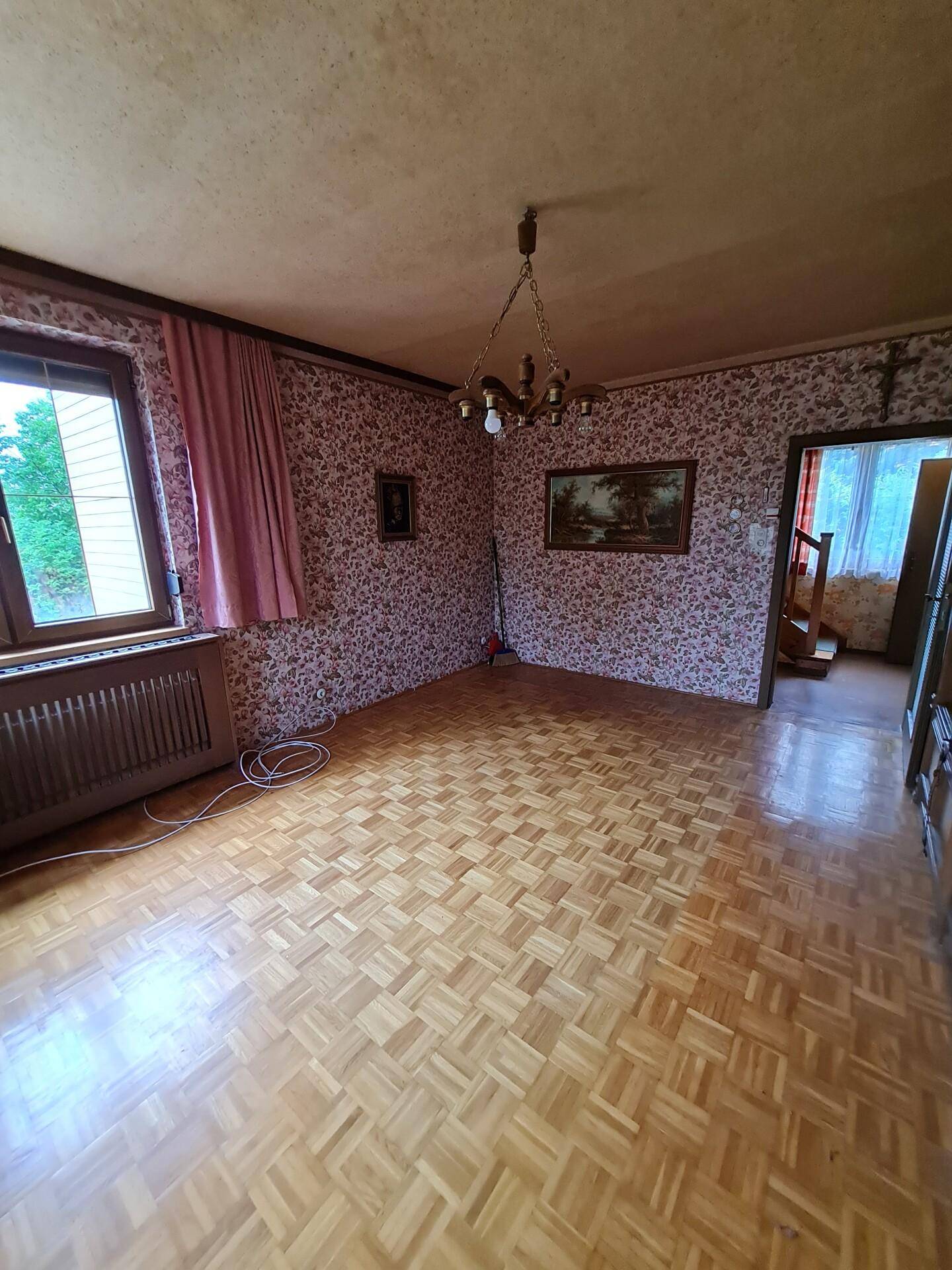 Leeres Wohnzimmer mit Parkettboden, floraler Tapete und Kronleuchter.