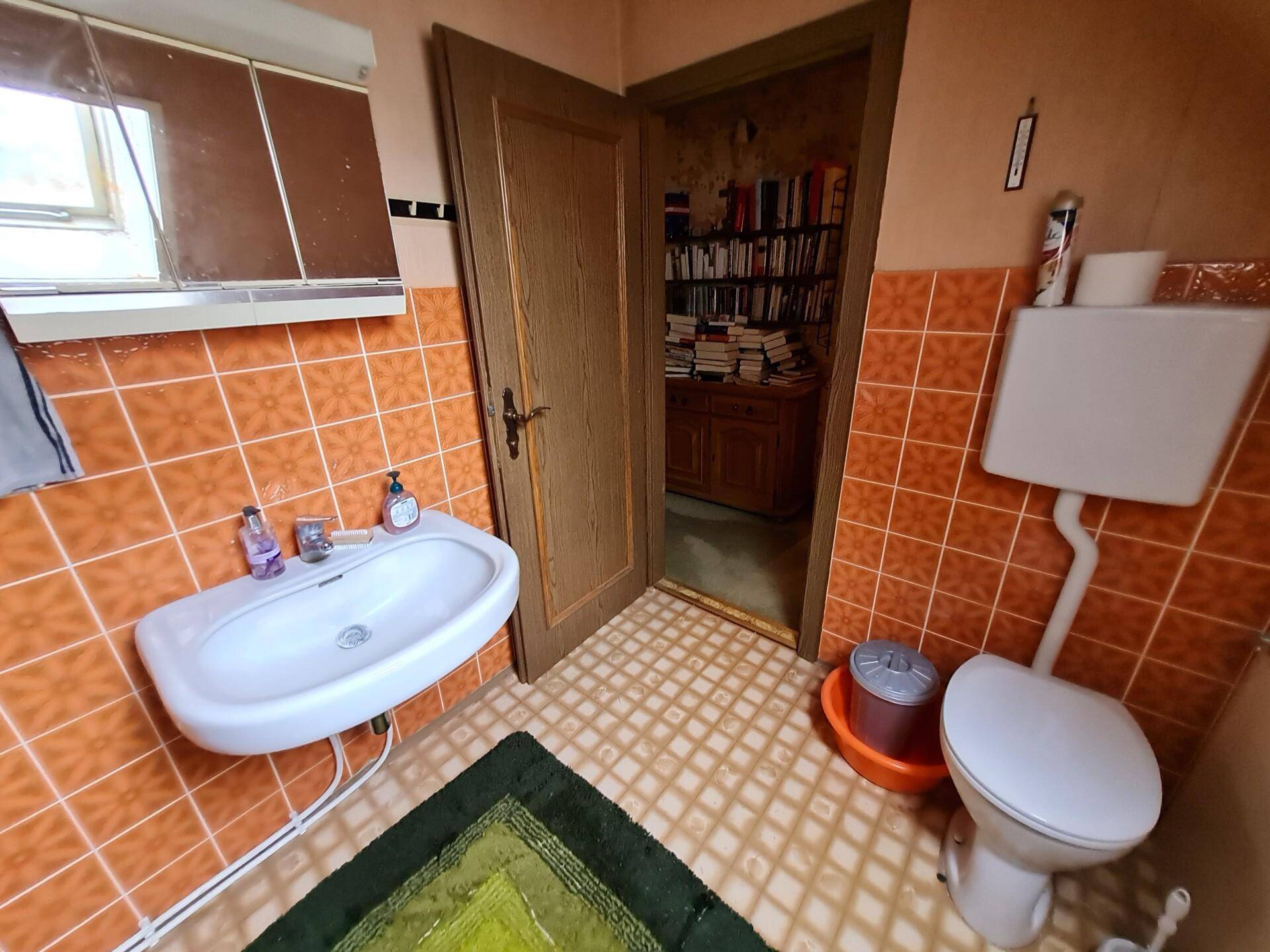 Badezimmer mit Waschbecken, WC und orangefarbenen Fliesen, mit Blick in einen Nebenraum.