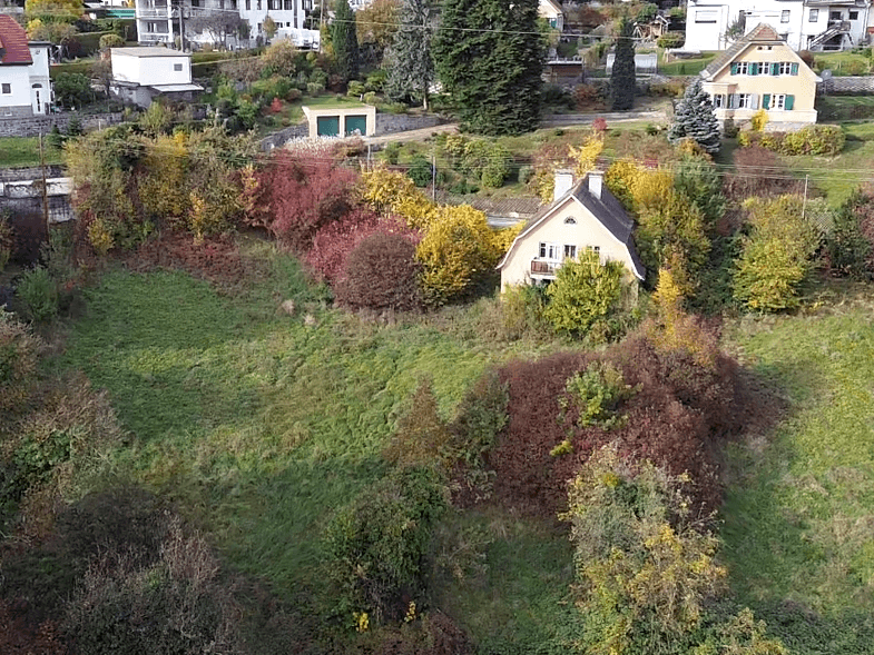 Nähere Luftaufnahme eines gelben Hauses mit kleinem Balkon, umgeben von herbstlicher Vegetation auf einem Hanggrundstück.
