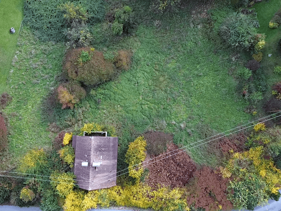 Senkrechte Luftaufnahme eines Gebäudes mit Flachdach, umgeben von herbstlicher Vegetation und einer angrenzenden Straße.