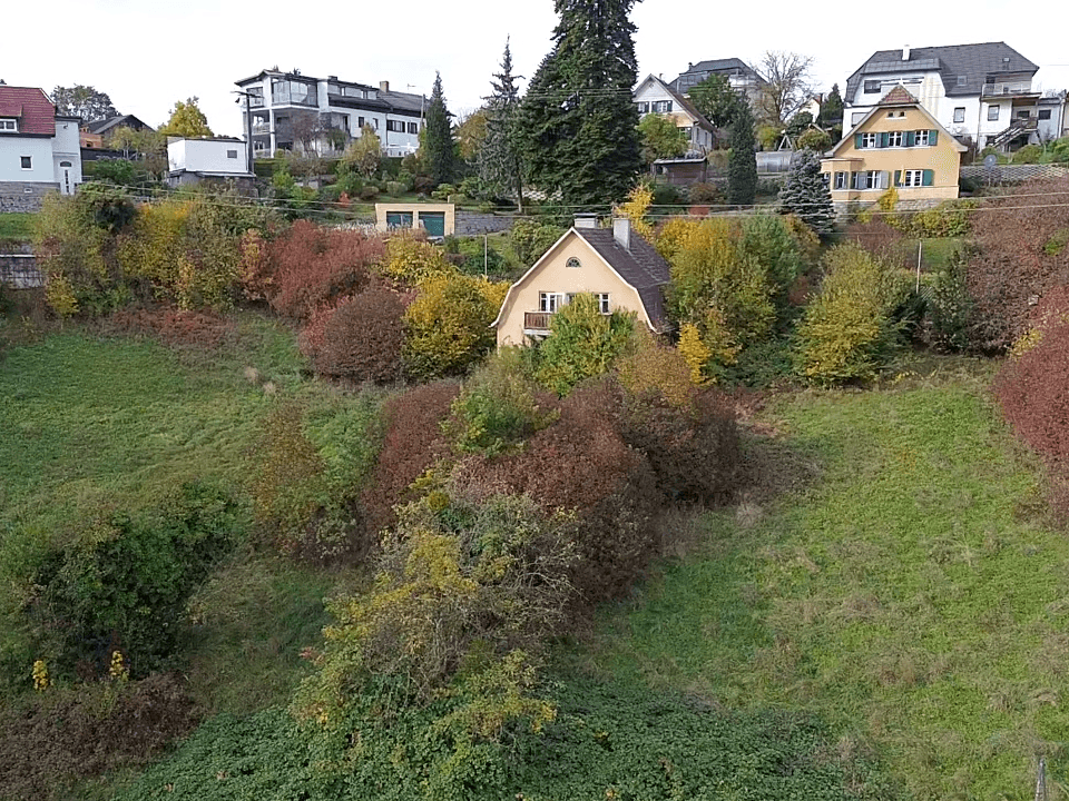 Luftaufnahme eines Hauses auf einem bewachsenen Hanggrundstück mit Herbstlaub und weiteren Häusern im Hintergrund.