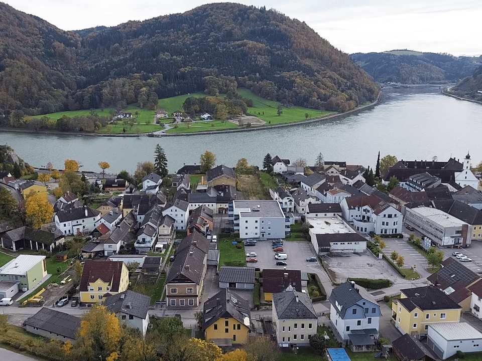 Panoramablick auf eine malerische Stadt am Flussufer, umgeben von bewaldeten Bergen und herbstlicher Landschaft.