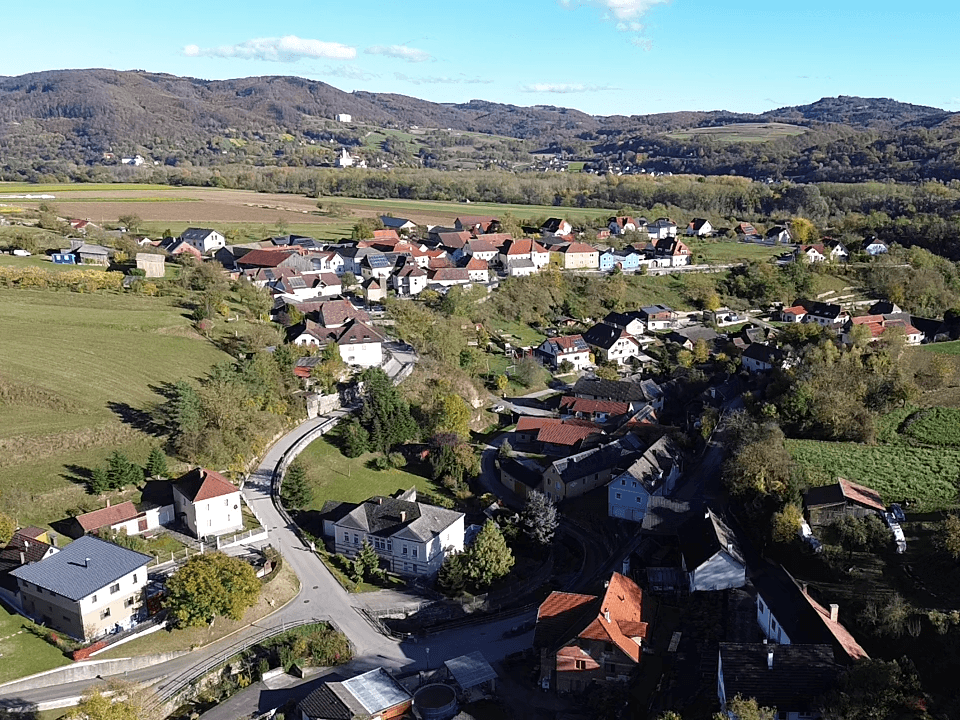 Panoramablick aus der Luft auf ein malerisches Dorf in einem Tal, umgeben von bewaldeten Hügeln und Feldern.