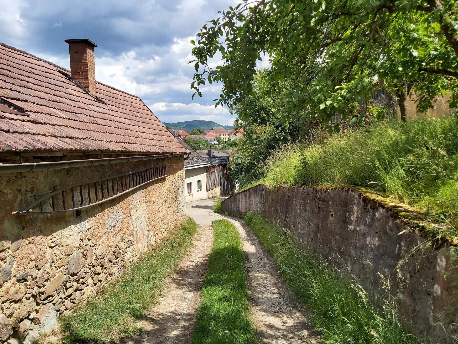 Schmaler Feldweg zwischen einer alten Steinmauer und einem traditionellen Haus mit Ziegeldach, umgeben von Grün.