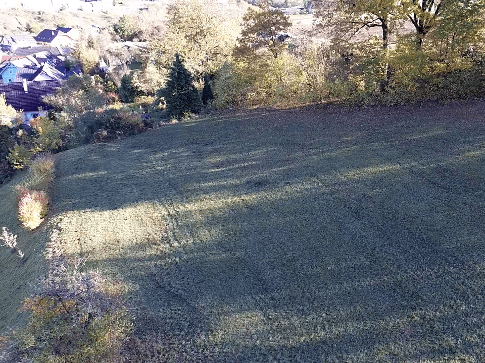 Luftaufnahme einer grünen Wiese mit langen Schatten, die zu einem Dorf in der Ferne abfällt.