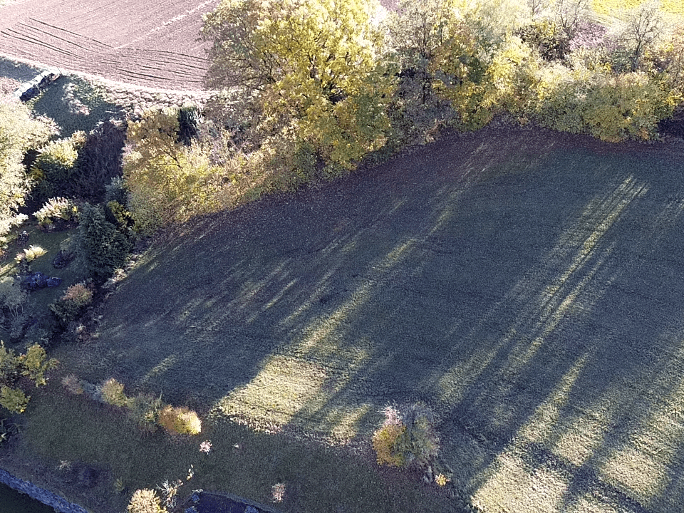 Luftaufnahme einer grünen Wiese, umgeben von Bäumen und landwirtschaftlichen Feldern, mit einem Dorf im Hintergrund.