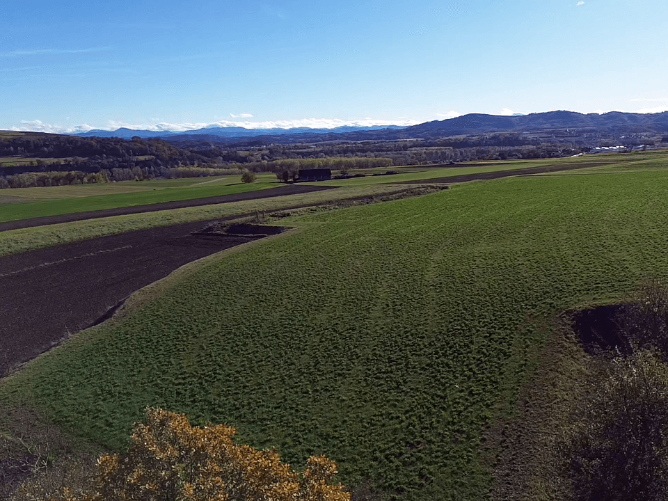 Luftaufnahme einer weiten Landschaft mit grünen Wiesen und braunen Feldern, umgeben von fernen Hügeln.