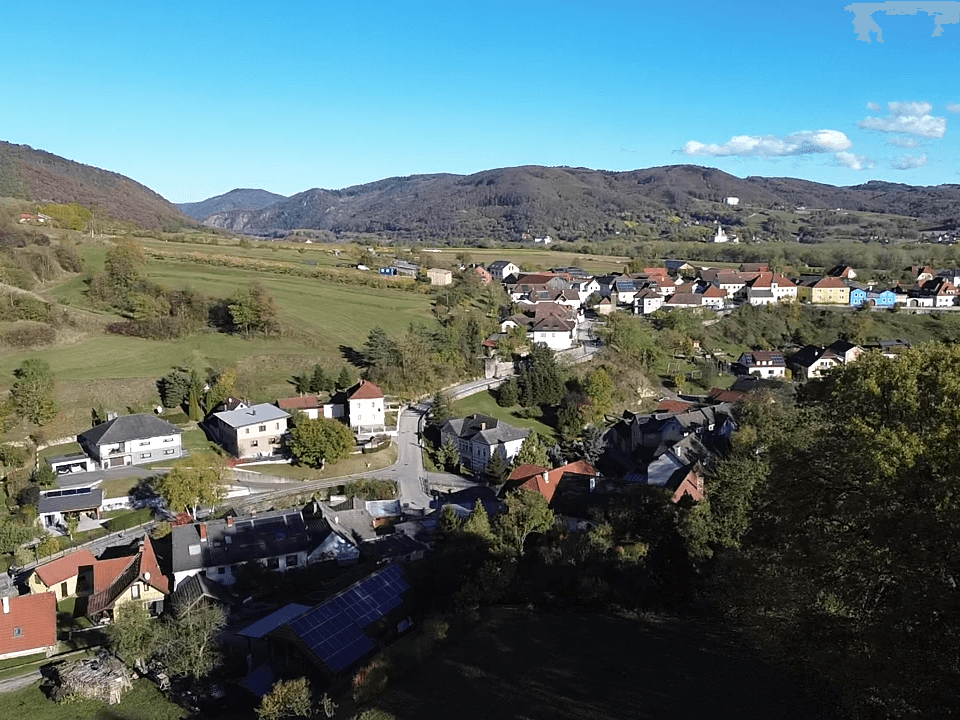 Panoramablick aus der Luft auf ein malerisches Dorf in einem Tal, umgeben von bewaldeten Hügeln und Feldern.