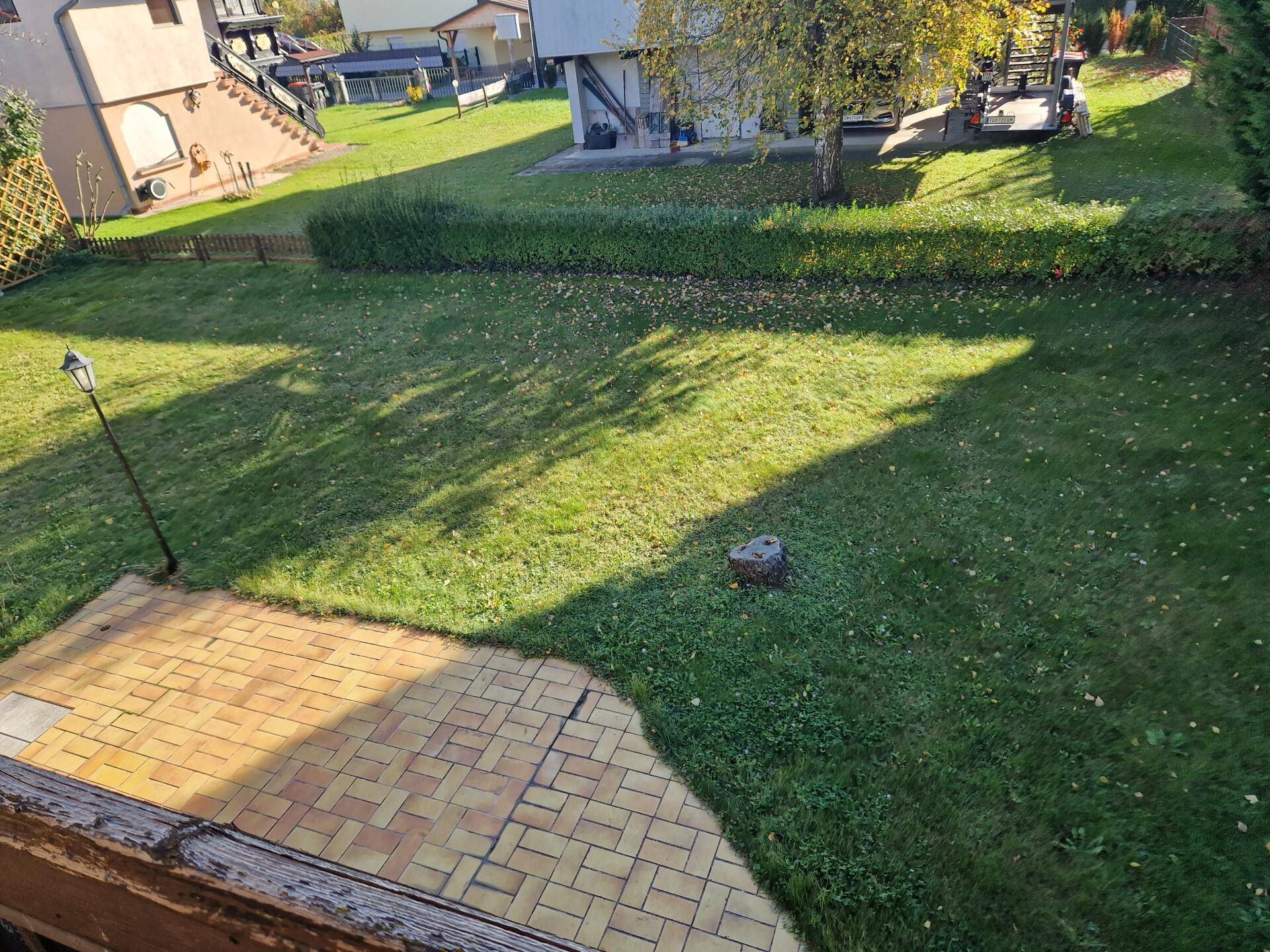 Sonniger Gartenbereich mit Rasenfläche und gepflasterter Terrasse, umgeben von Hecken.