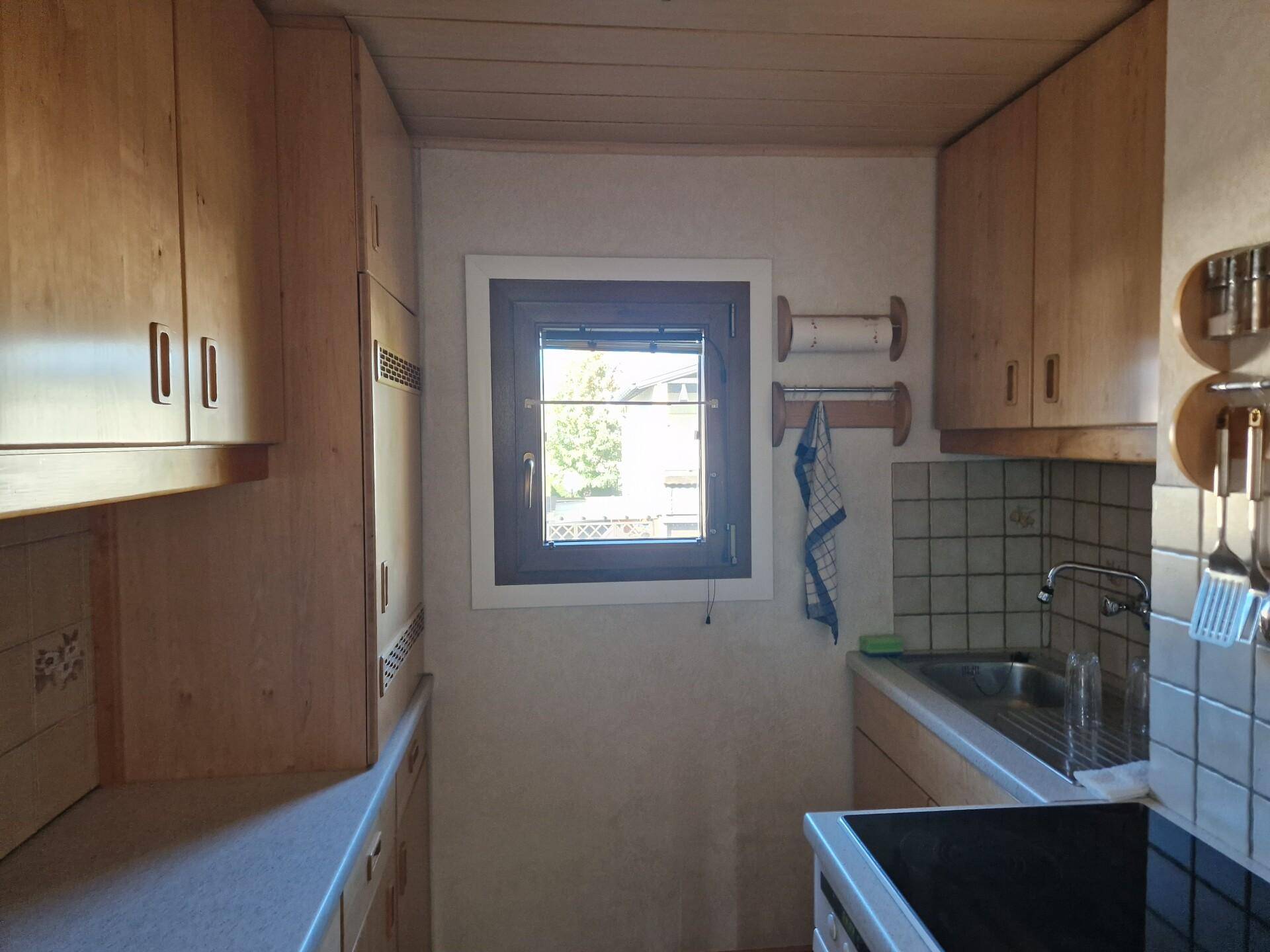 Funktionale Küche mit Holzschränken, Fliesenboden und Fenster mit Blick ins Grüne.