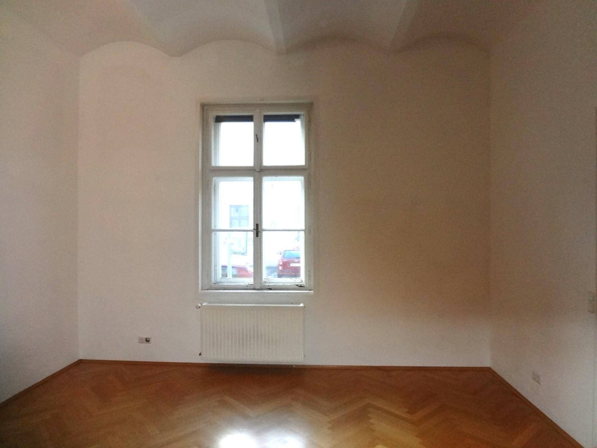 Leerer Raum mit großem Fenster, Heizkörper und klassischem Fischgrätparkettboden.