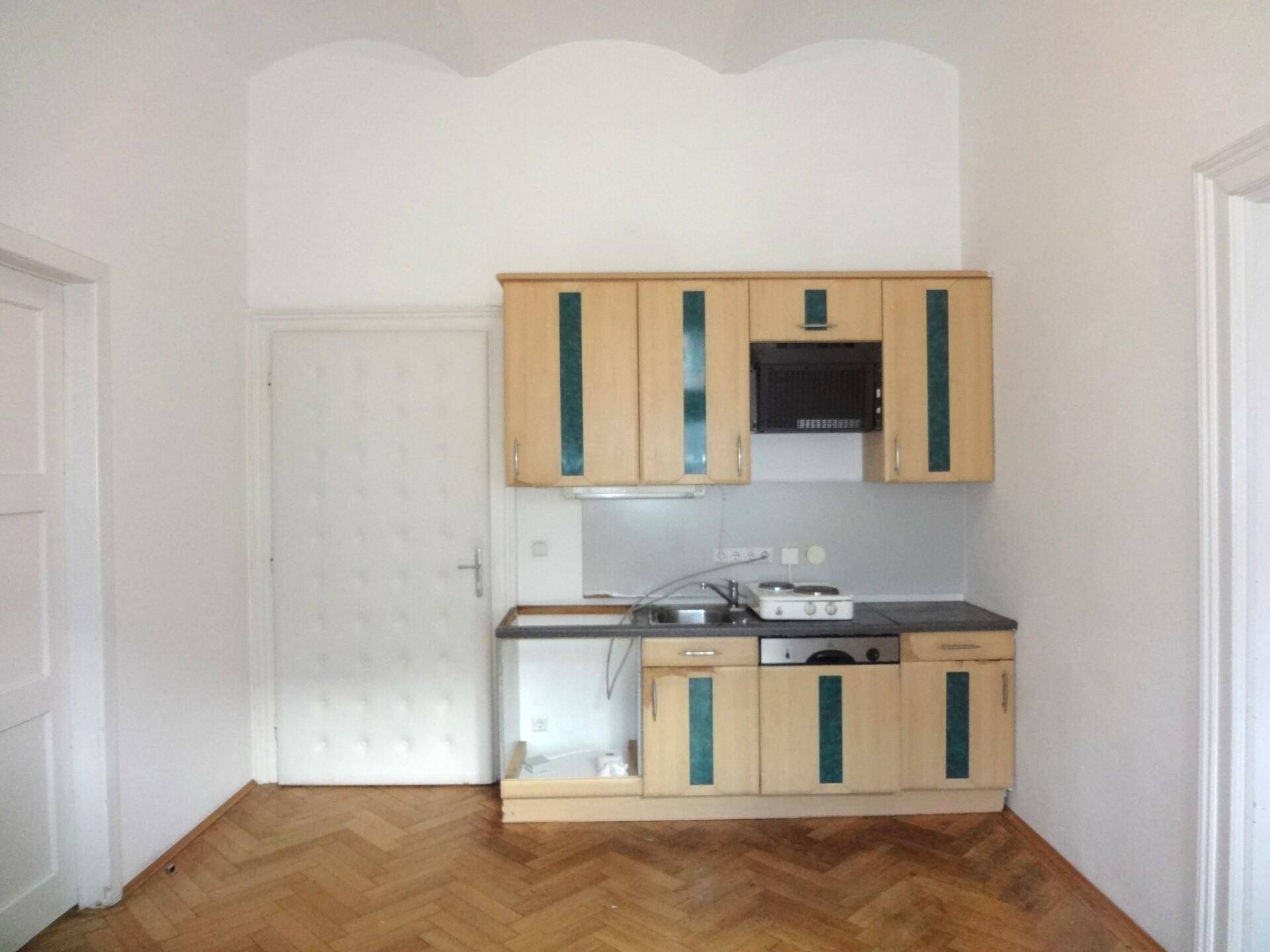 Kleine Küchenzeile mit hellen Holzschränken, Spüle und Kochfeld, ergänzt durch Fischgrätparkett.