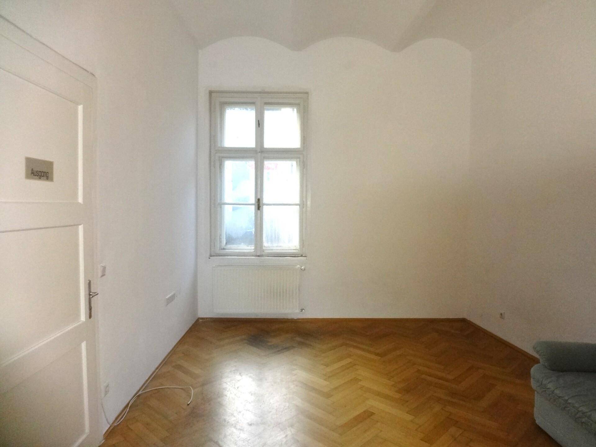 Heller, leerer Raum mit einem großen Fenster und glänzendem Fischgrätparkettboden.