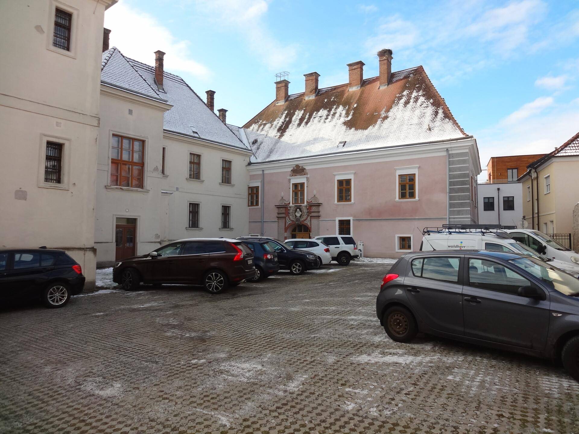 Innenhof mit geparkten Autos und historischen Gebäuden im Hintergrund, leicht verschneit.