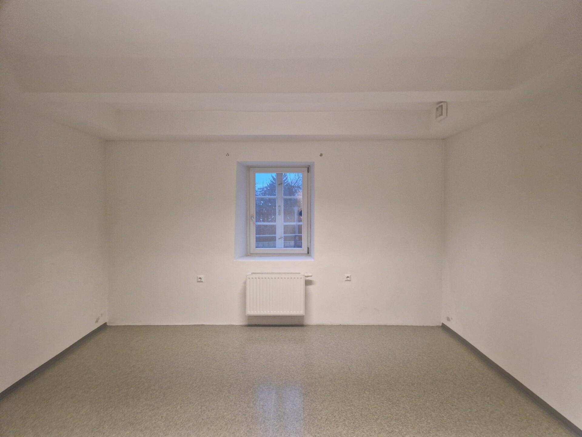 Heller, leerer Raum mit einem Fenster, Heizkörper und Linoleumboden, ideal für Büro oder Lager.