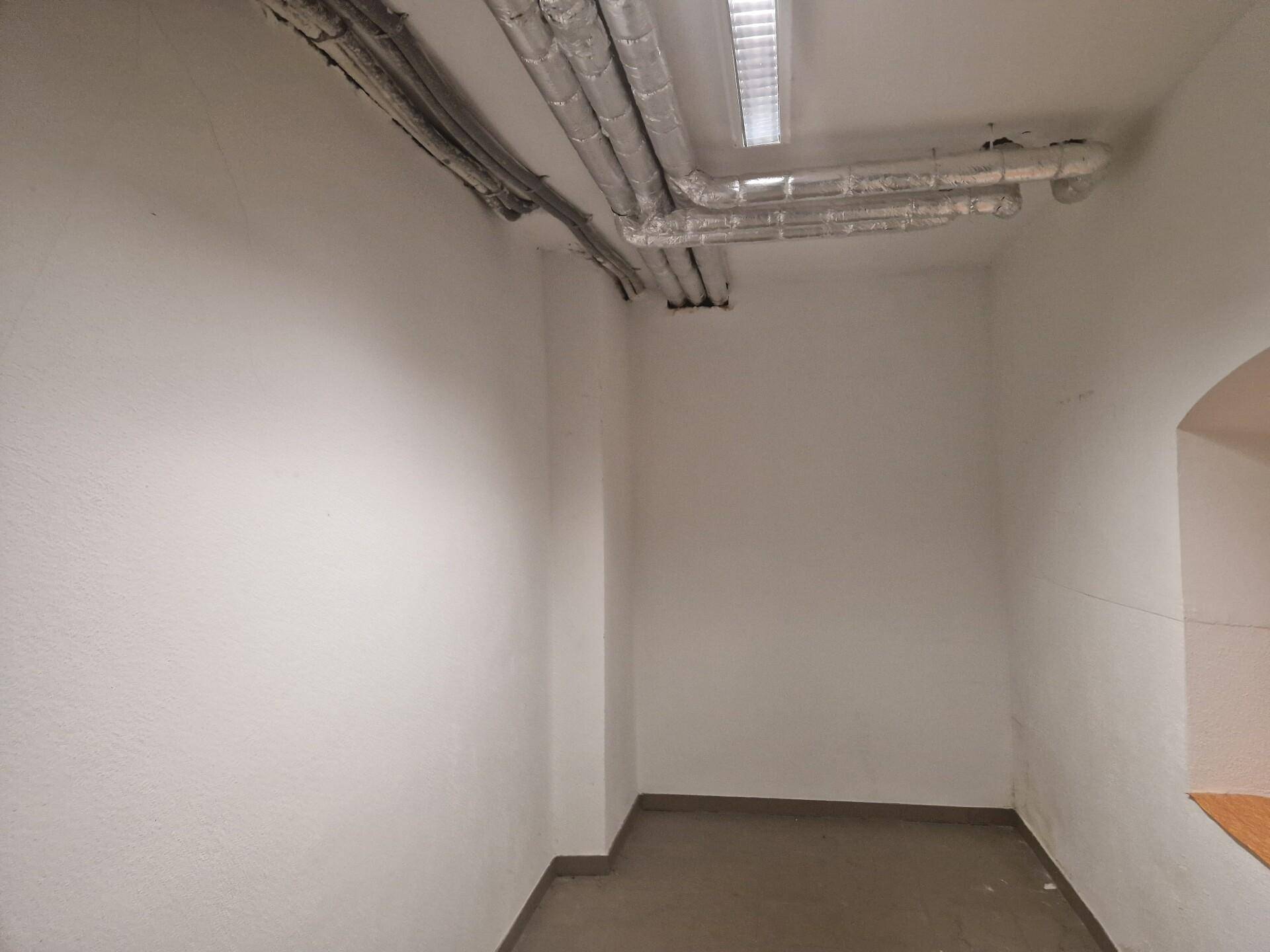 Abstellraum mit sichtbaren Rohren an der Decke und einfachen weißen Wänden, gefliester Boden.