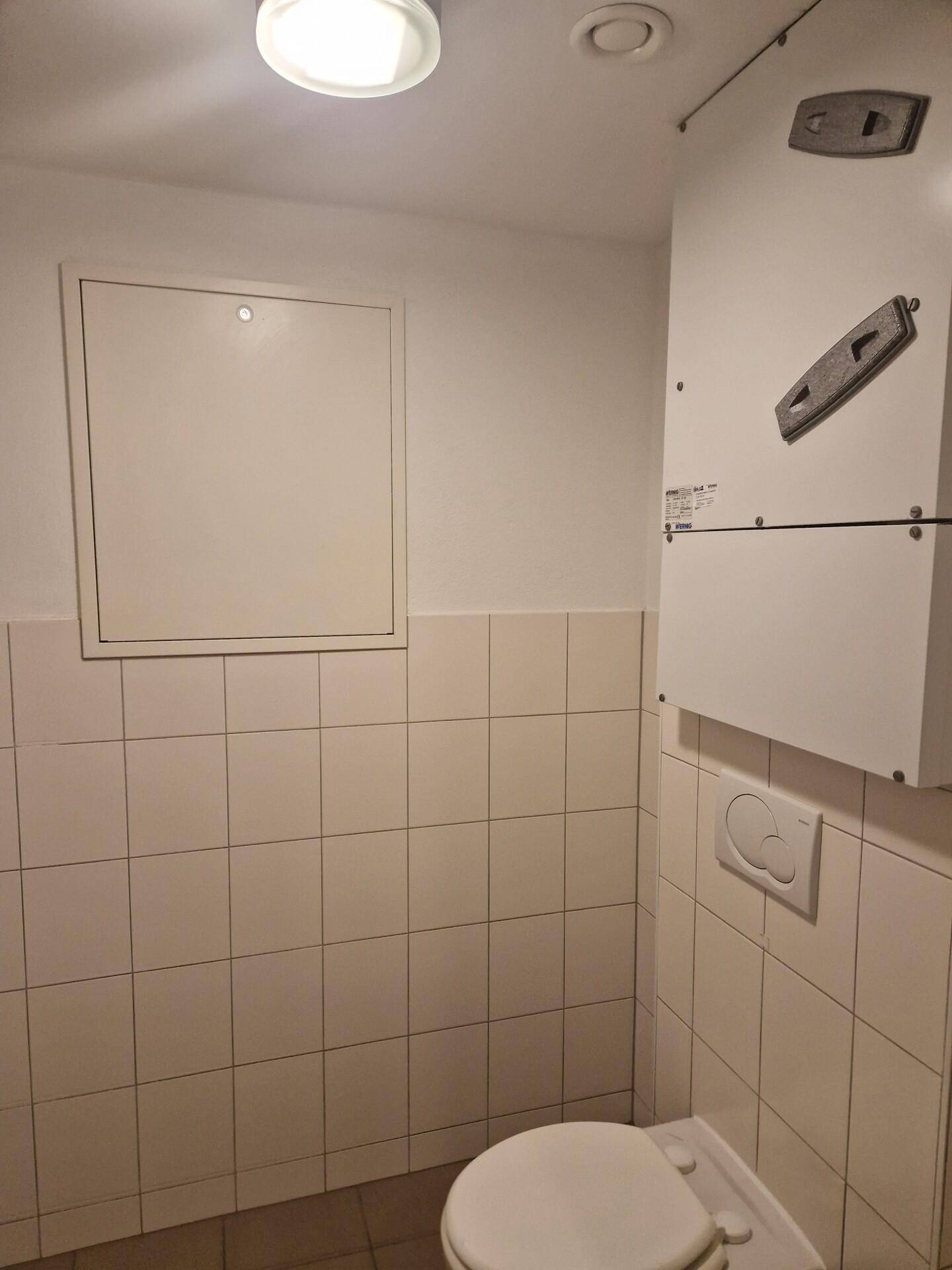 Kleine Toilette mit weißen Fliesen und einem Spülkasten, mit Deckenleuchte und Lüftung.