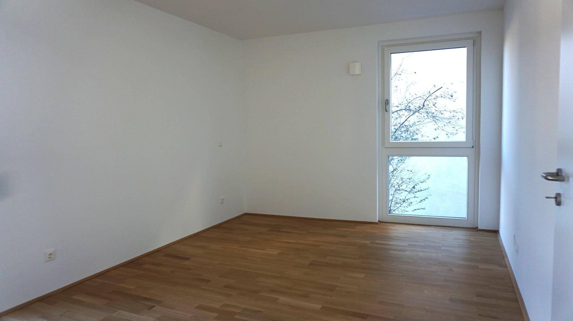 Helles Zimmer mit Parkettboden und einem großen Fenster, das viel Tageslicht hereinlässt.