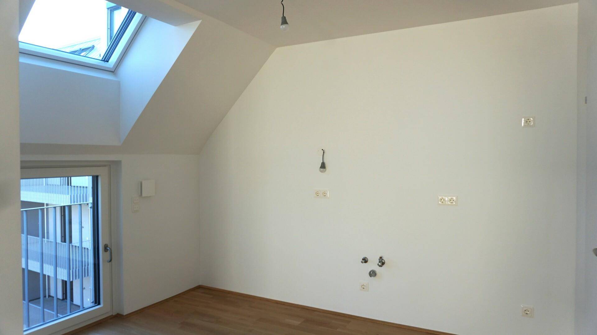 Leerer Dachgeschossraum mit Dachfenster und vorbereiteten Anschlüssen für eine Küche oder Bad.
