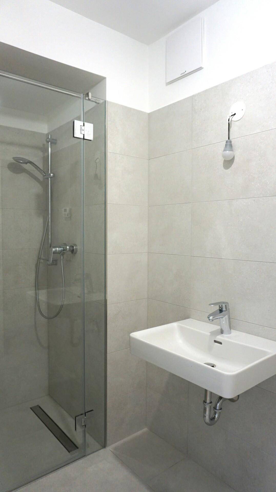 Helles Badezimmer mit moderner Duschkabine, Waschbecken und grauen Wandfliesen.