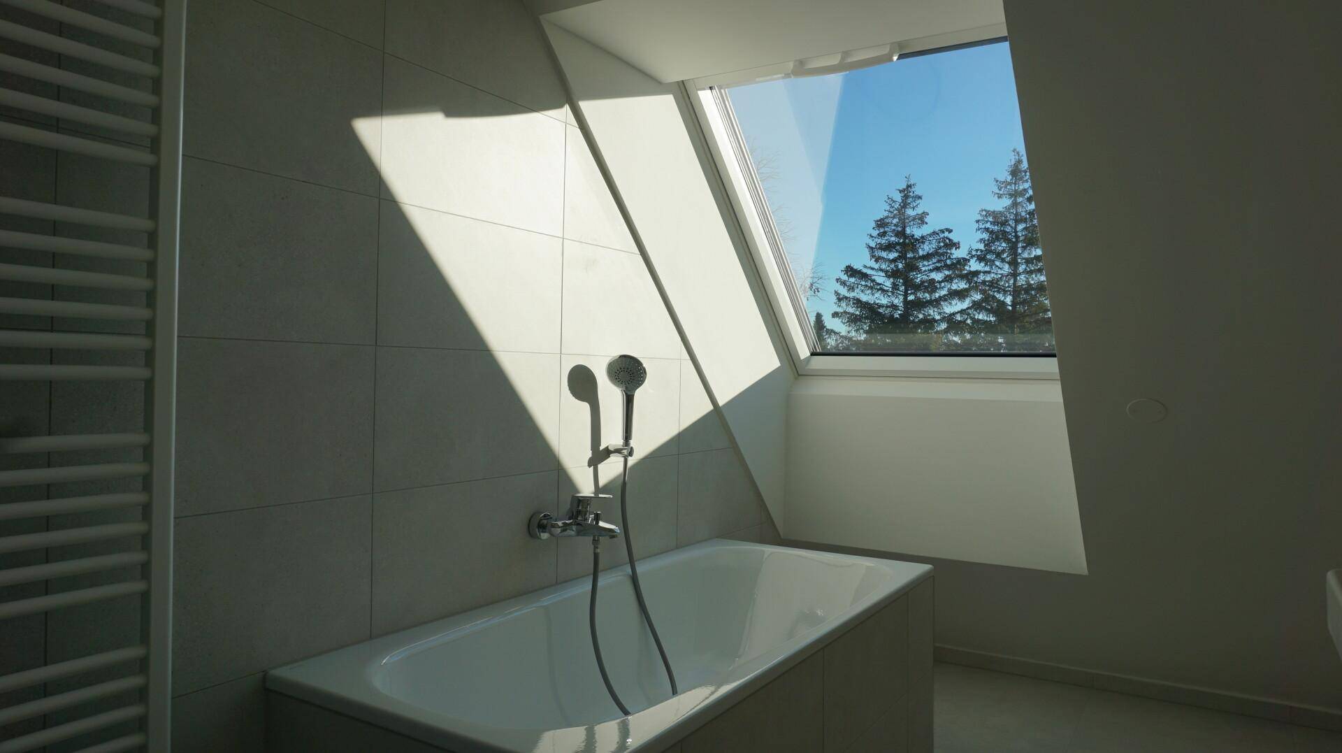 Sonnenlichtdurchflutetes Badezimmer mit Badewanne, Duscharmatur und Blick ins Grüne durch das Dachfenster.