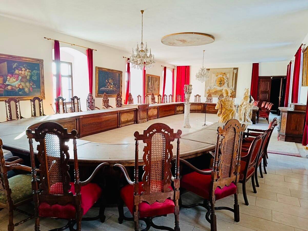 Prunkvoller Speisesaal mit einem sehr langen Tisch, vielen Stühlen und großen Fenstern mit roten Vorhängen.