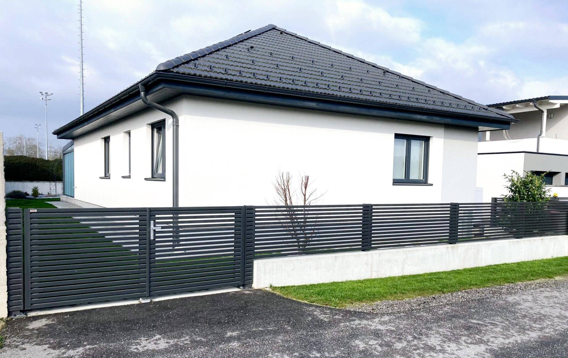 Moderne Hausfassade mit dunklem Dach und stilvollem Zaun im Vorgartenbereich.