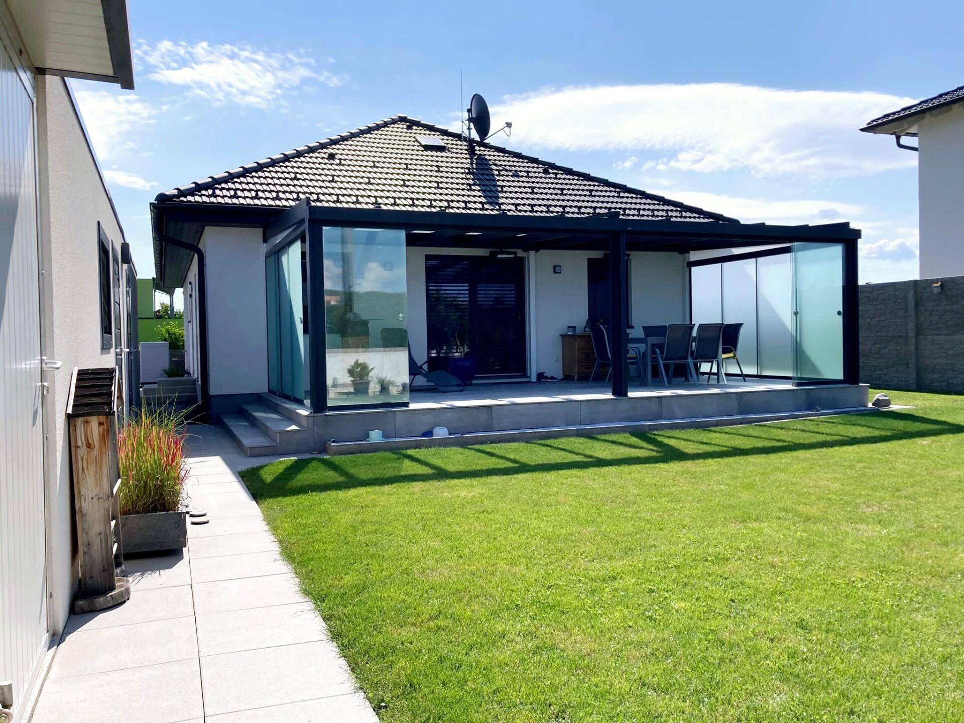 Sonniger Blick auf das moderne Haus mit großem Wintergarten und gepflegtem Rasen im Garten.