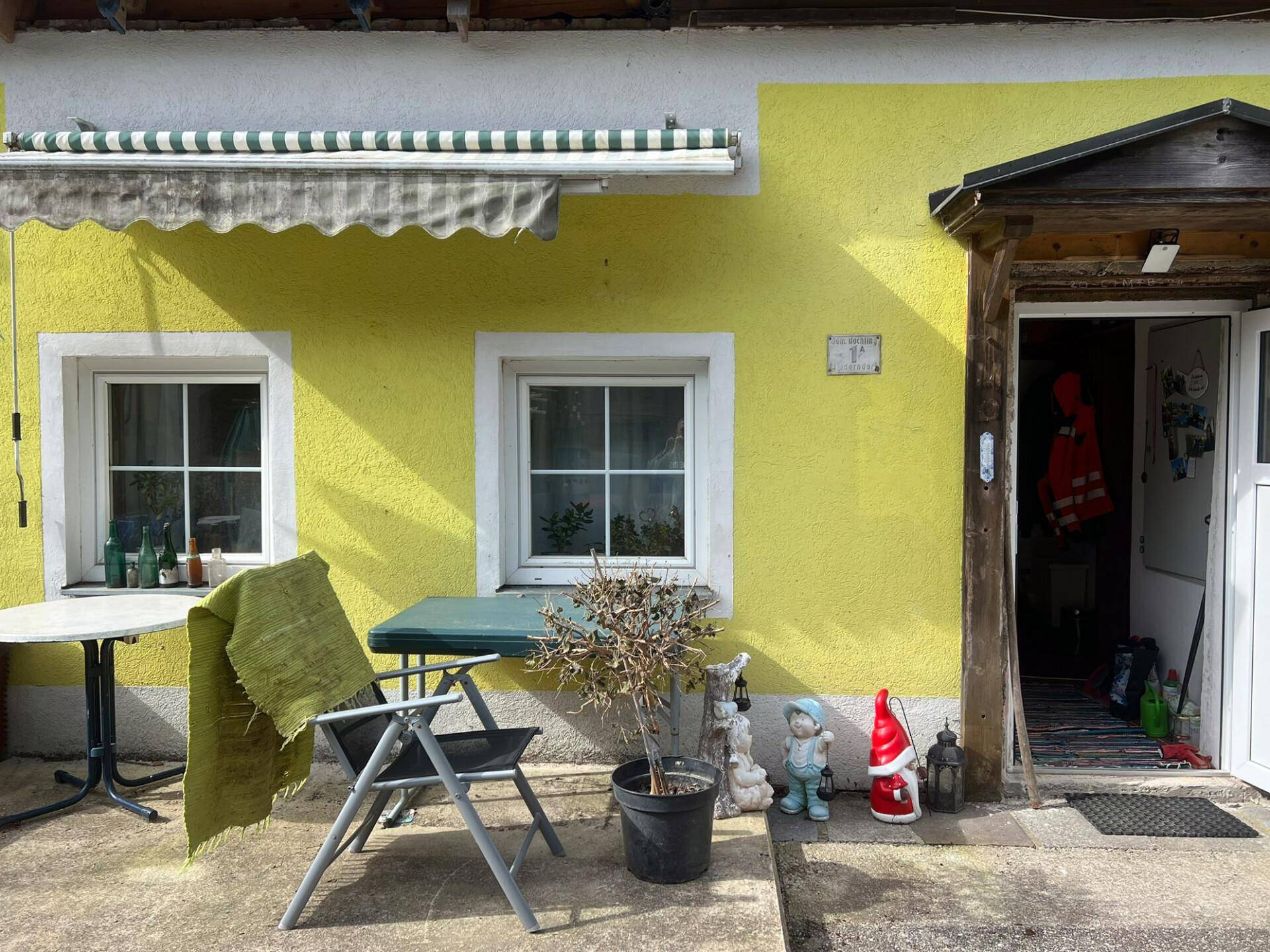 Nahaufnahme der gelben Hausfassade mit einem kleinen Sitzbereich und Gartenzwergen am Eingang.
