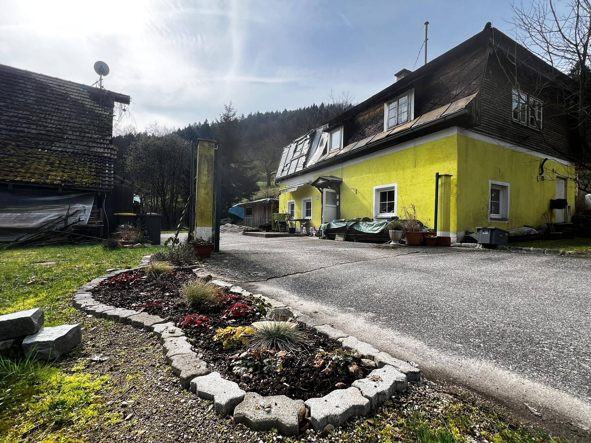 Ansicht des gelben Hauses mit Zufahrt und einem kleinen Blumenbeet im Vordergrund.