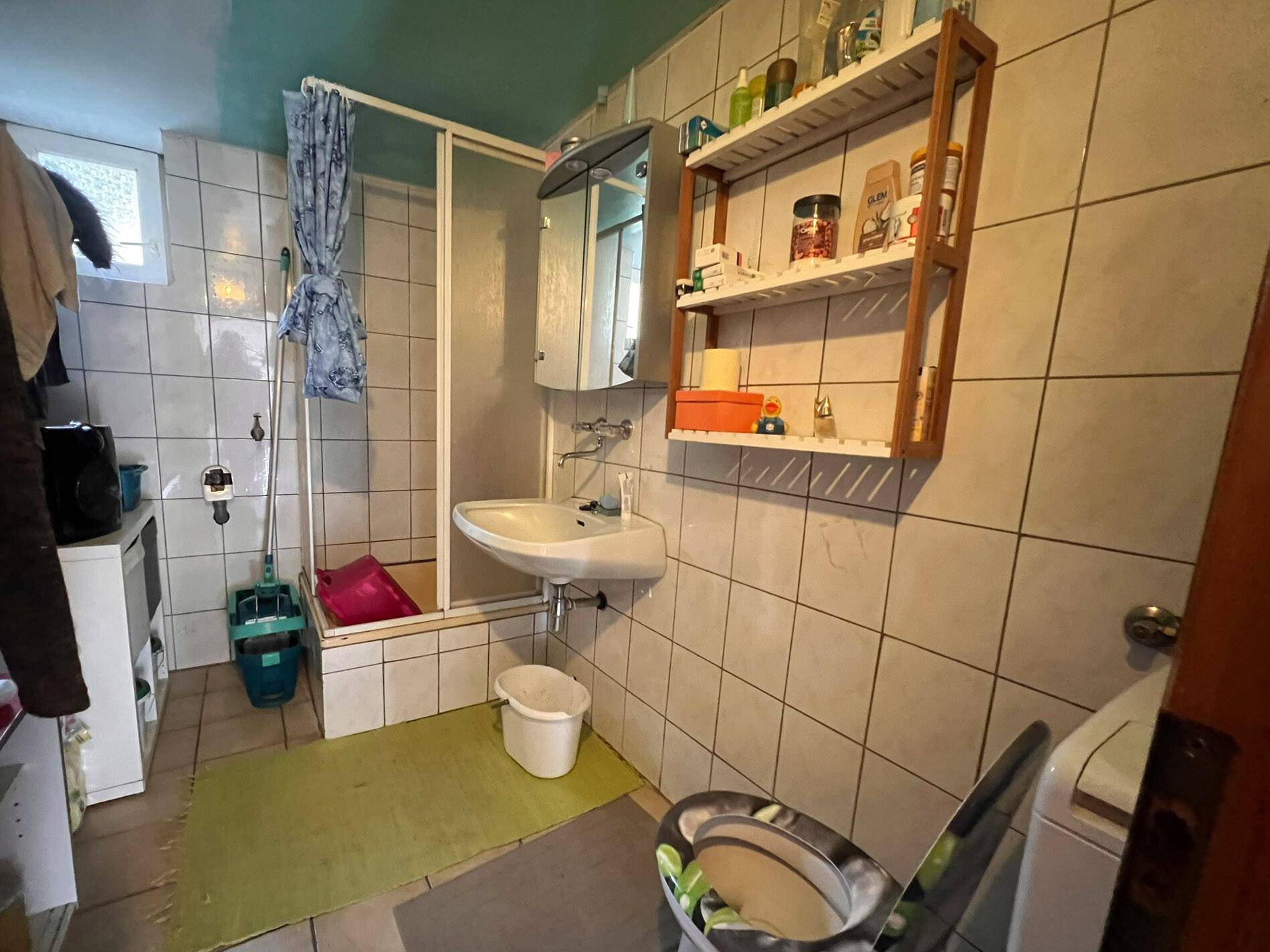 Badezimmer mit weißen Fliesen, Duschkabine, Waschbecken und Toilette in älterem Zustand.