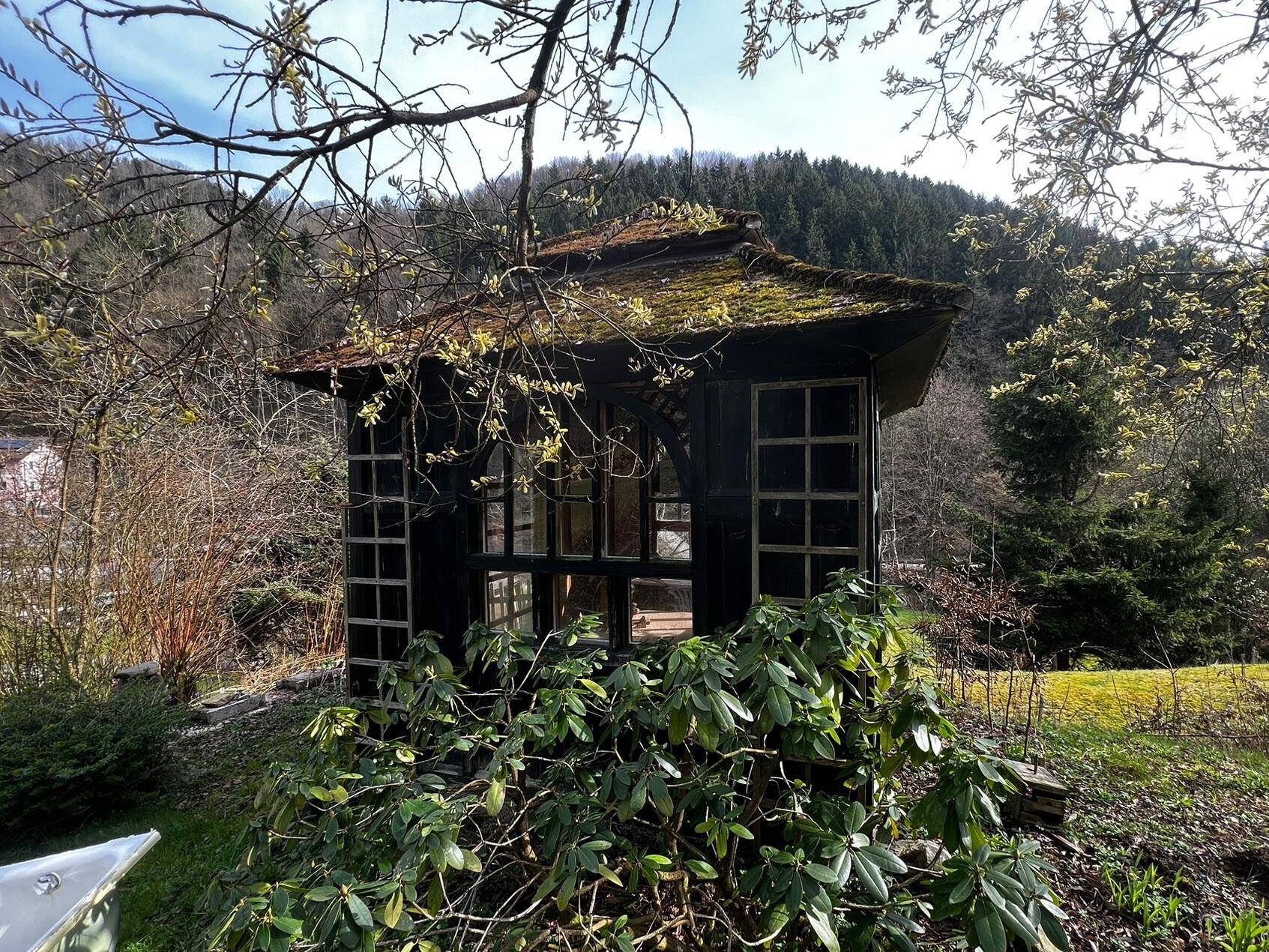 Kleiner, rustikaler Holzpavillon in einer grünen Gartenanlage, umgeben von Bäumen.