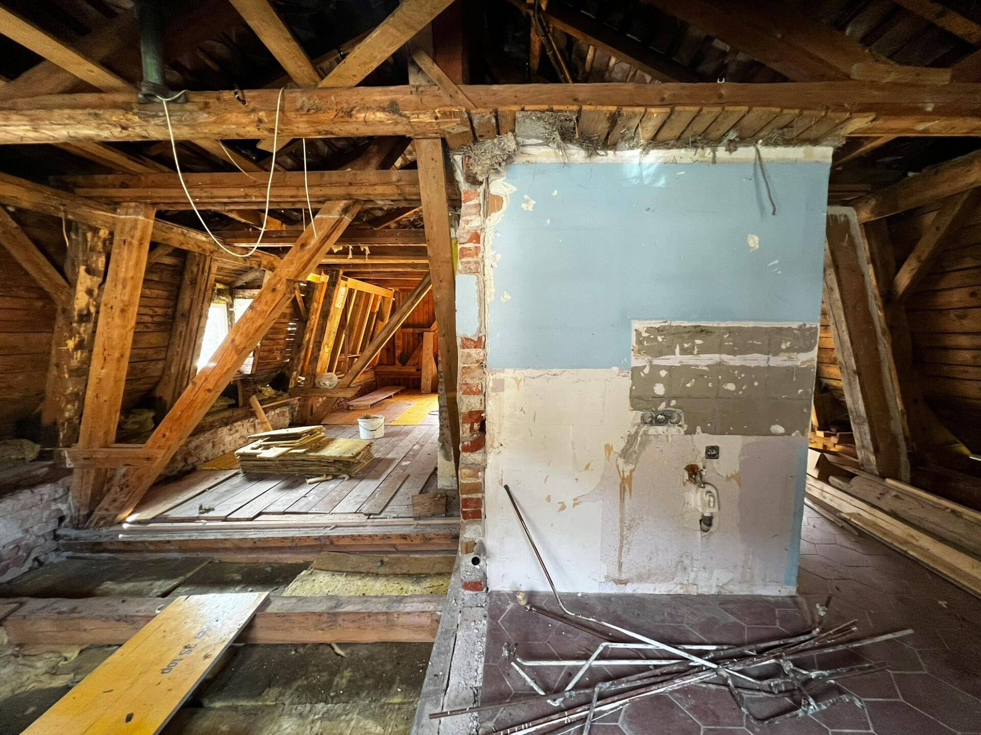 Dachboden im Umbau mit freiliegenden Holzbalken, teilweise verputzter Wand und Baumaterialien.