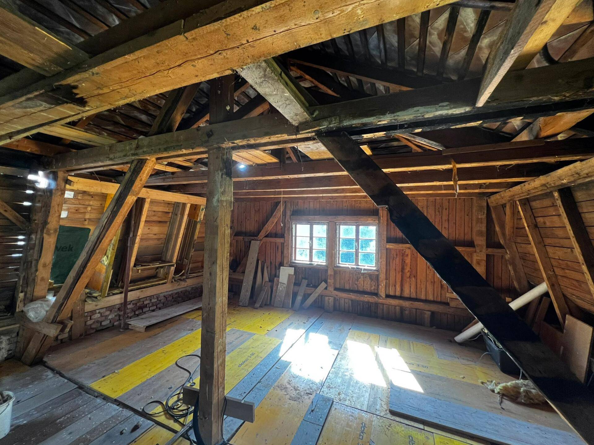 Dachboden im Umbau mit freiliegenden Holzbalken, Fenster und neuer Bodenisolierung.