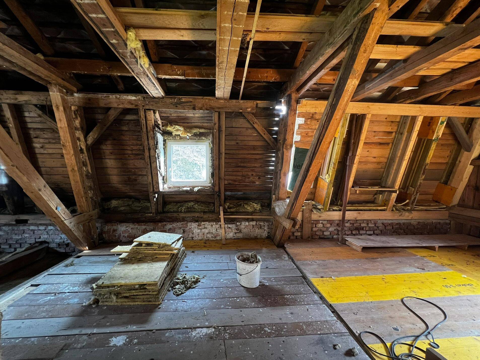 Dachboden im Umbau mit freiliegenden Holzbalken, Fenster und altem Bodenbelag.