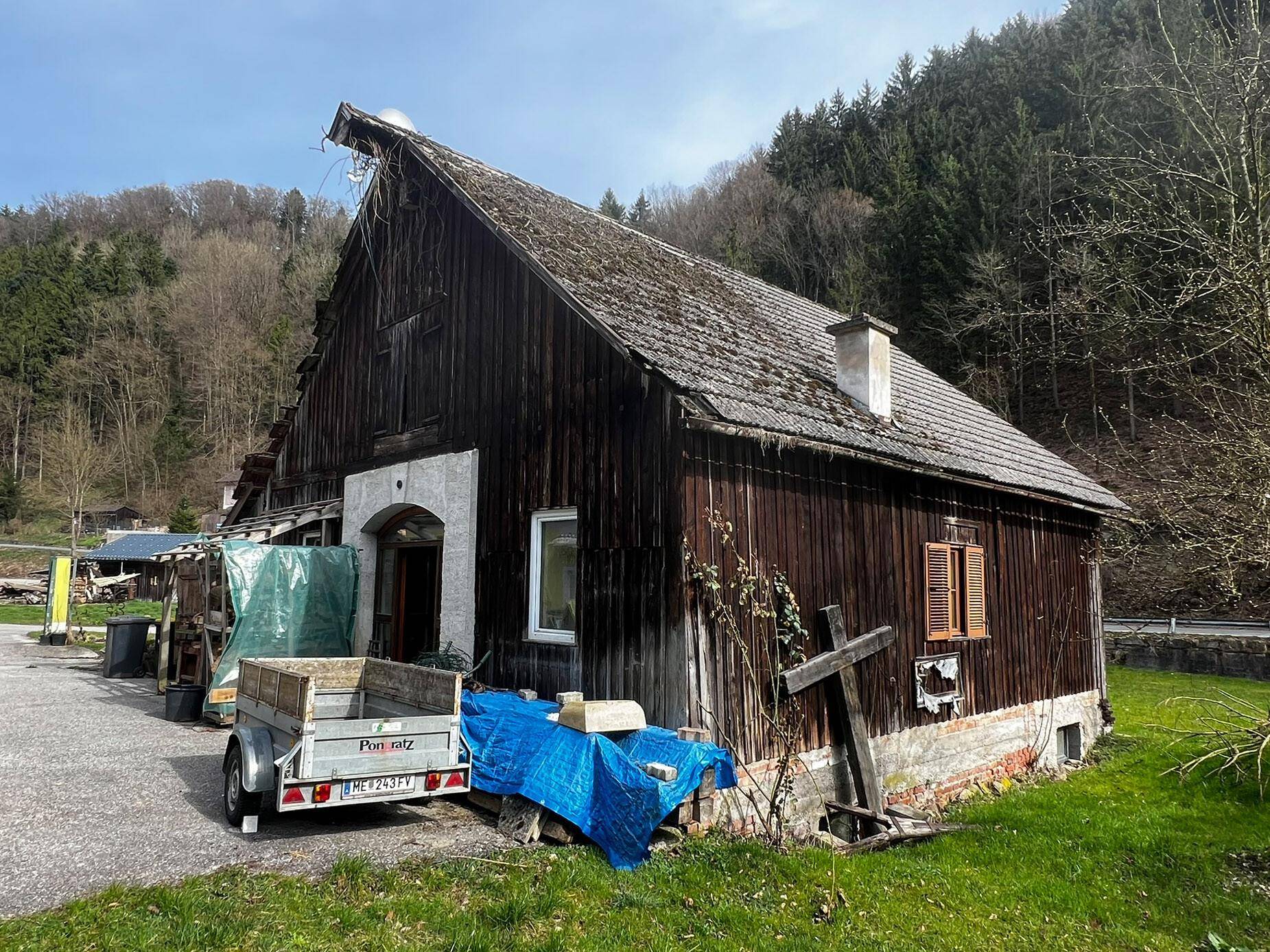 Altes Holzgebäude mit moosbewachsenem Dach in ländlicher Umgebung, umgeben von Bäumen.