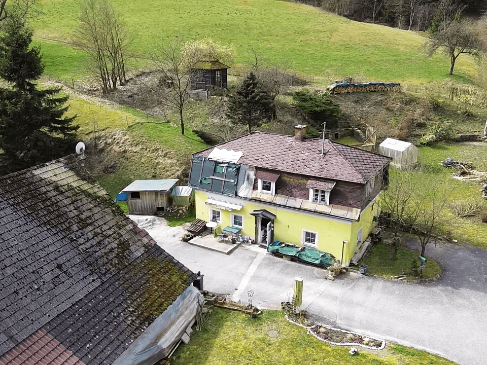 Vogelperspektive des gelben Hauses mit umliegenden Grünflächen und bewaldeten Hügeln.