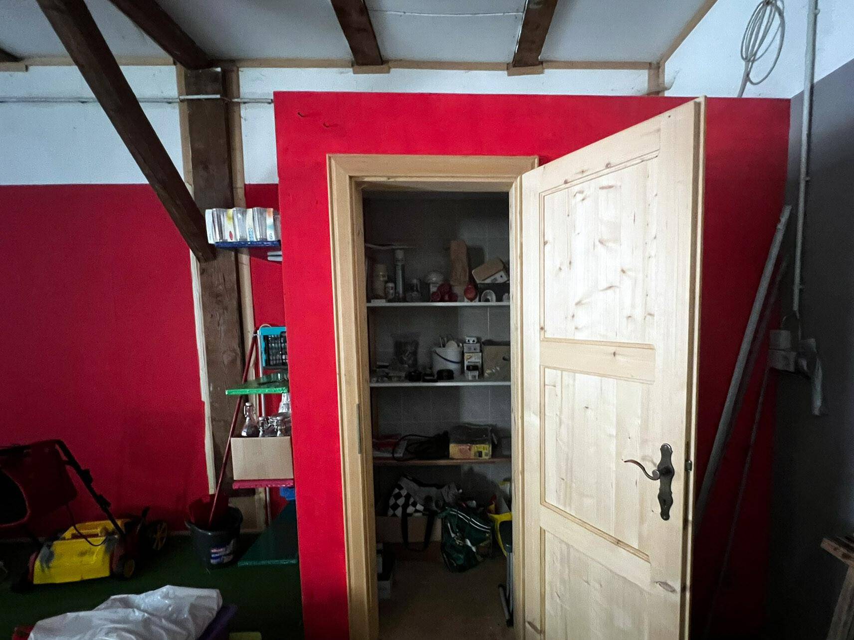 Kleiner Abstellraum mit Regalen, zugänglich durch eine Holztür in einem rot gestrichenen Bereich.