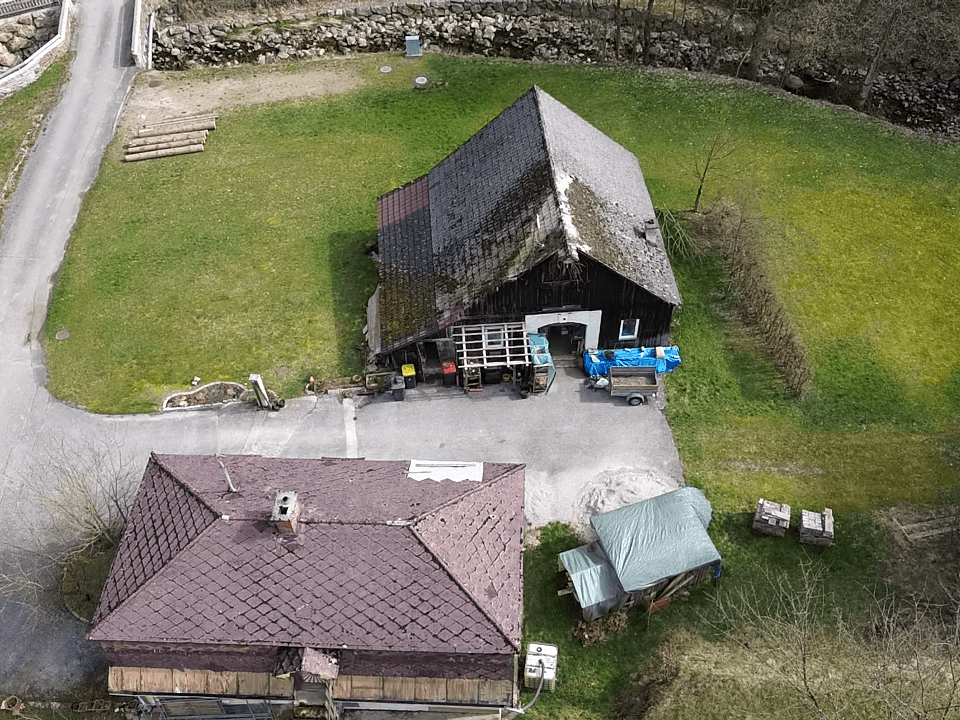 Luftaufnahme des Anwesens mit Holzscheune und Teilen des gelben Hauses, umgeben von Natur.