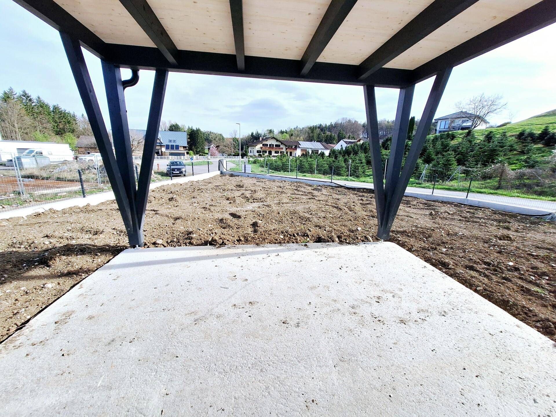 Unfertige Terrasse mit Betonboden und Holzüberdachung, umgeben von Erde und Blick ins Grüne.