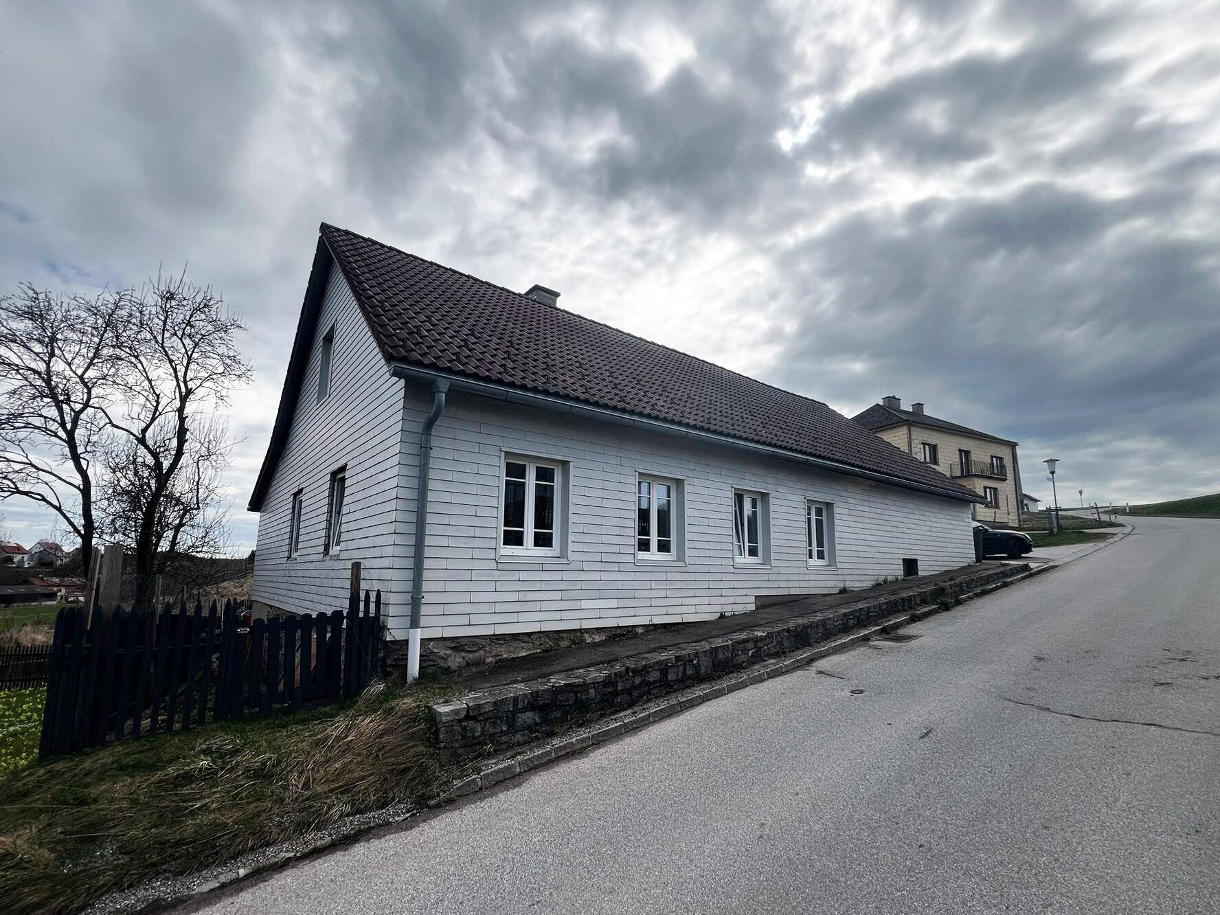 Seitenansicht eines Hauses mit heller Fassade und Satteldach unter bewölktem Himmel.