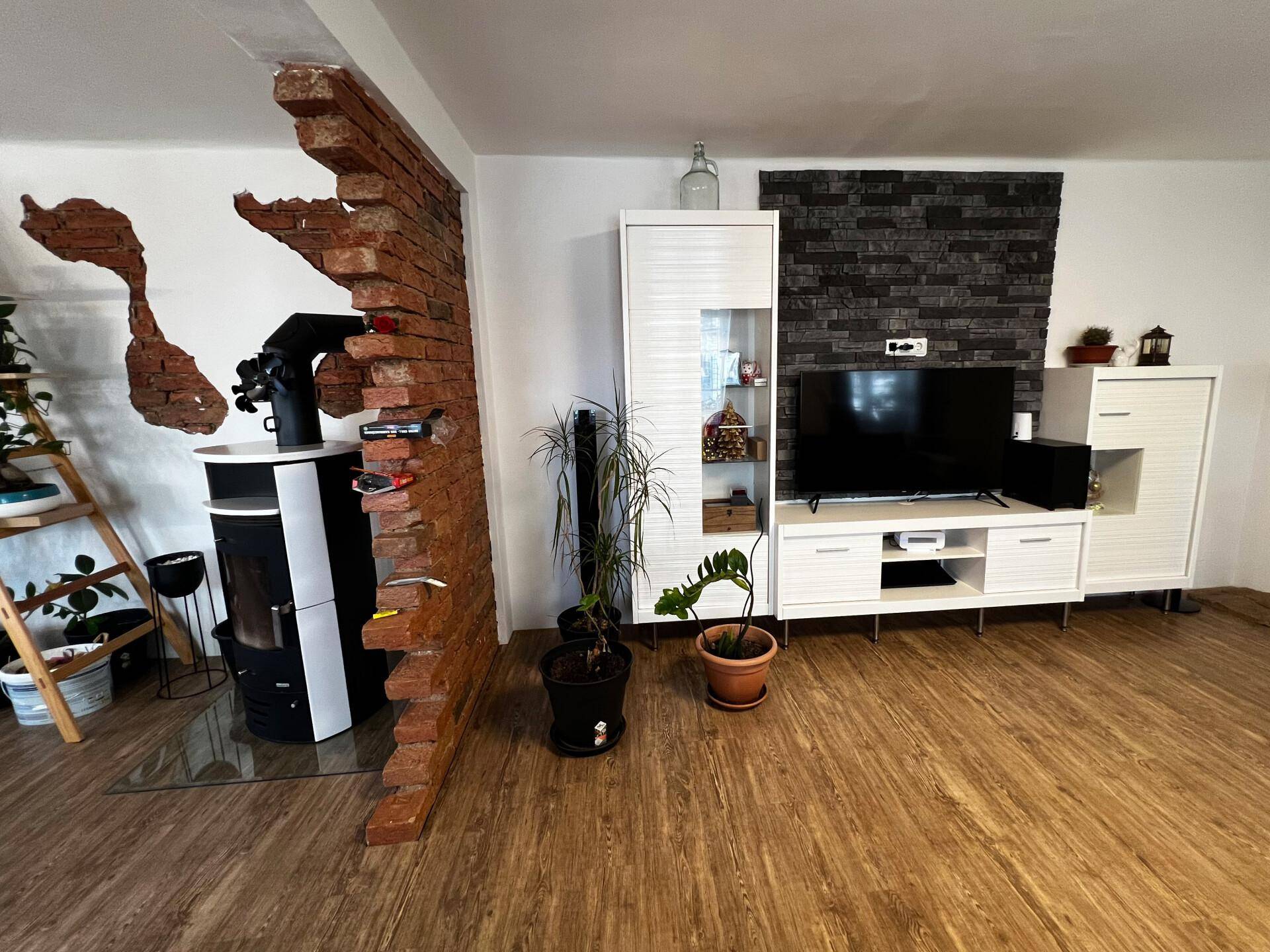 Wohnzimmer mit TV-Möbeln, Pflanzen und einem modernen Holzofen, sichtbar durch eine Ziegelwandöffnung.