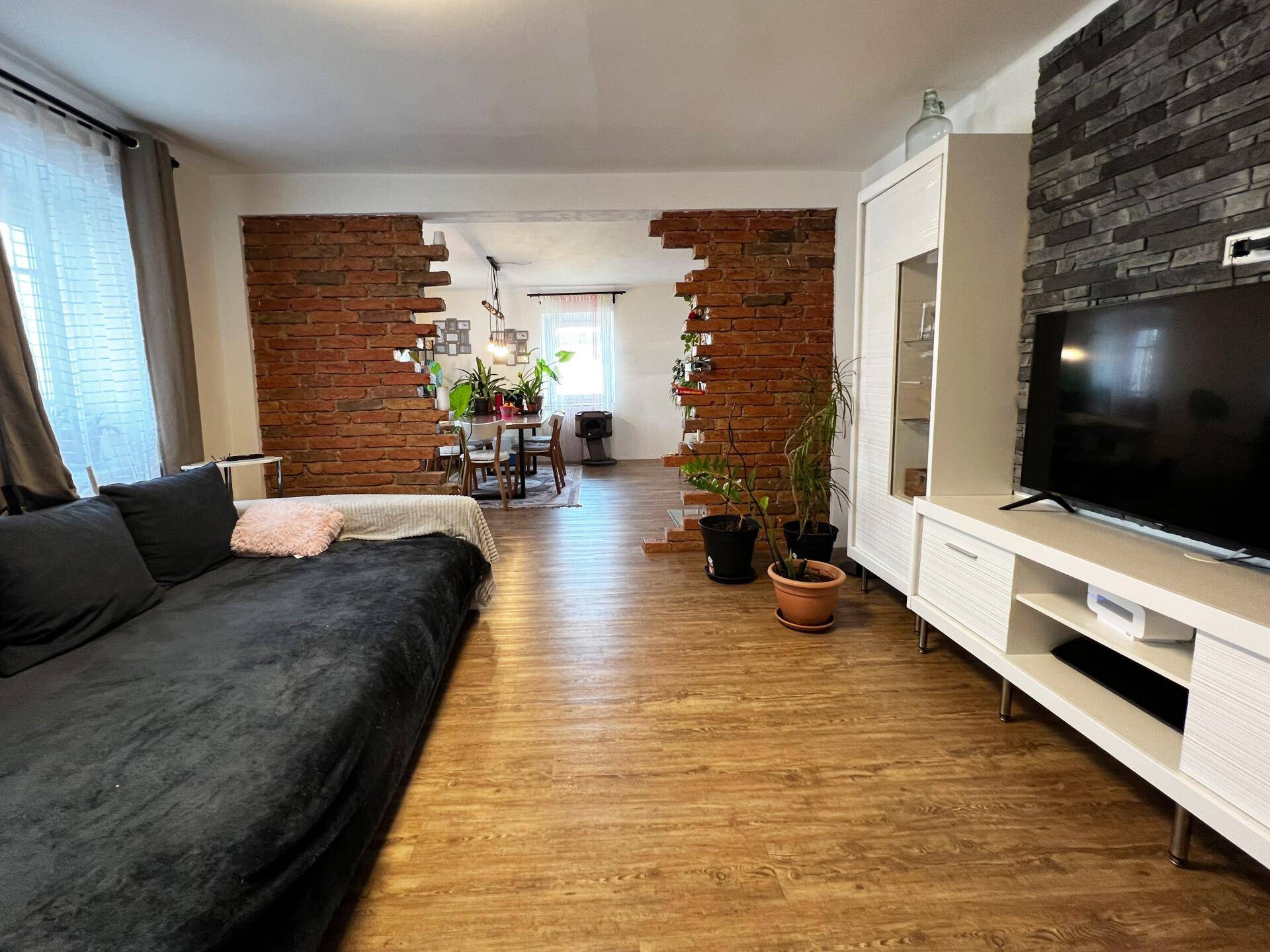 Geräumiger Wohnbereich mit Sofa, TV-Möbeln und offenem Zugang zum Esszimmer durch eine Ziegelwand.