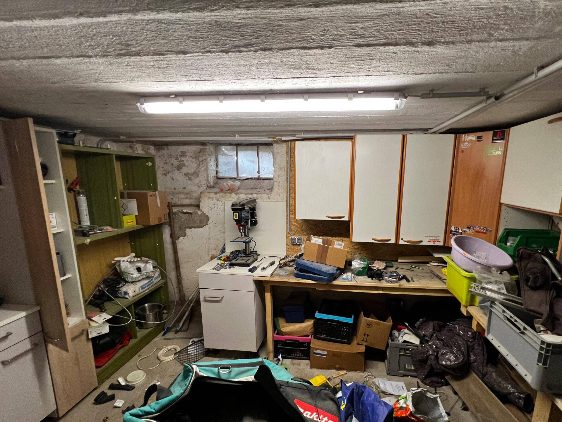 Werkstatt im Keller mit Werkbank, Regalen und verschiedenen Werkzeugen und Materialien.