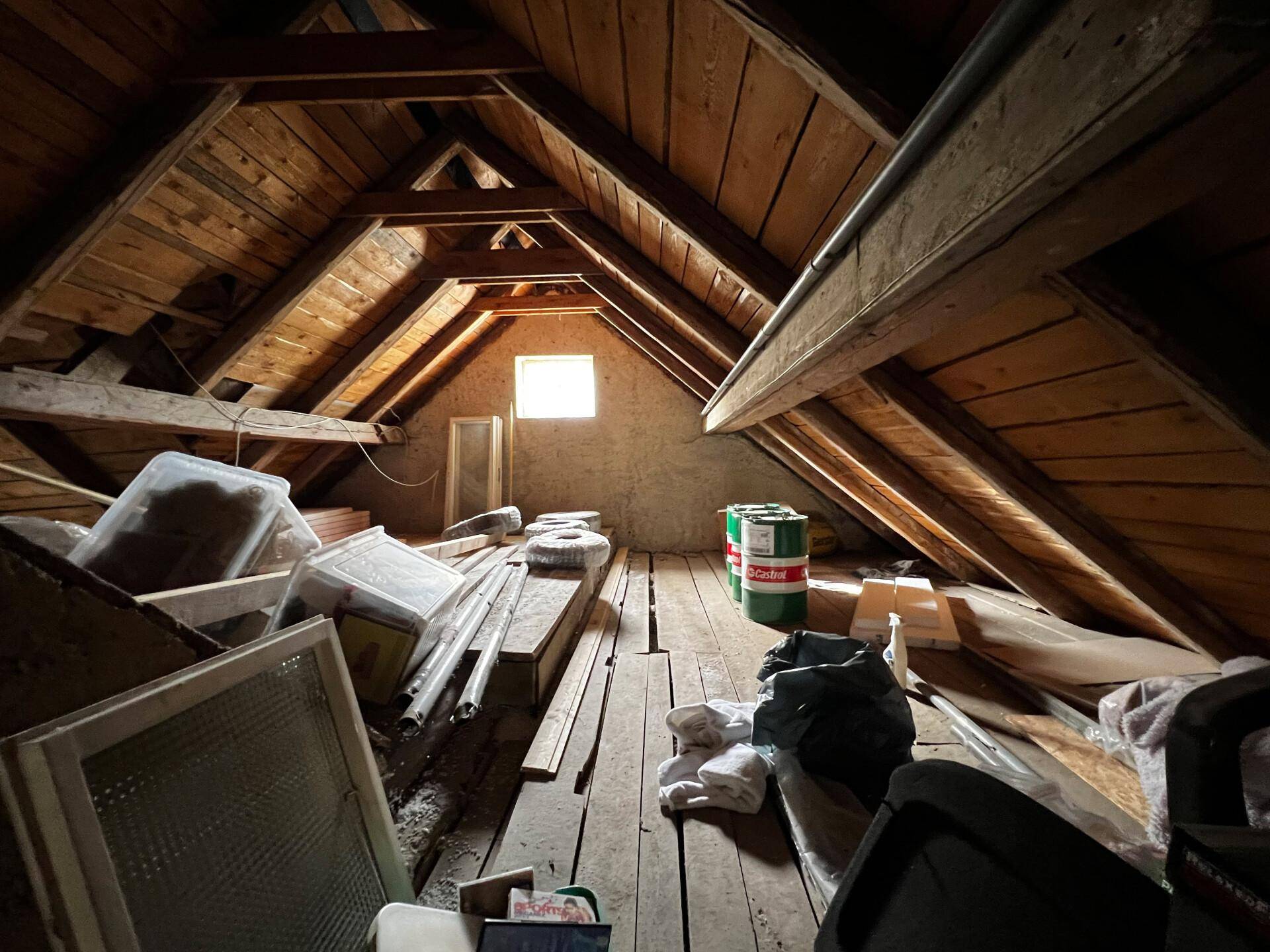 Unausgebauter Dachboden mit sichtbaren Holzbalken und einem kleinen Fenster, genutzt als Lagerraum.