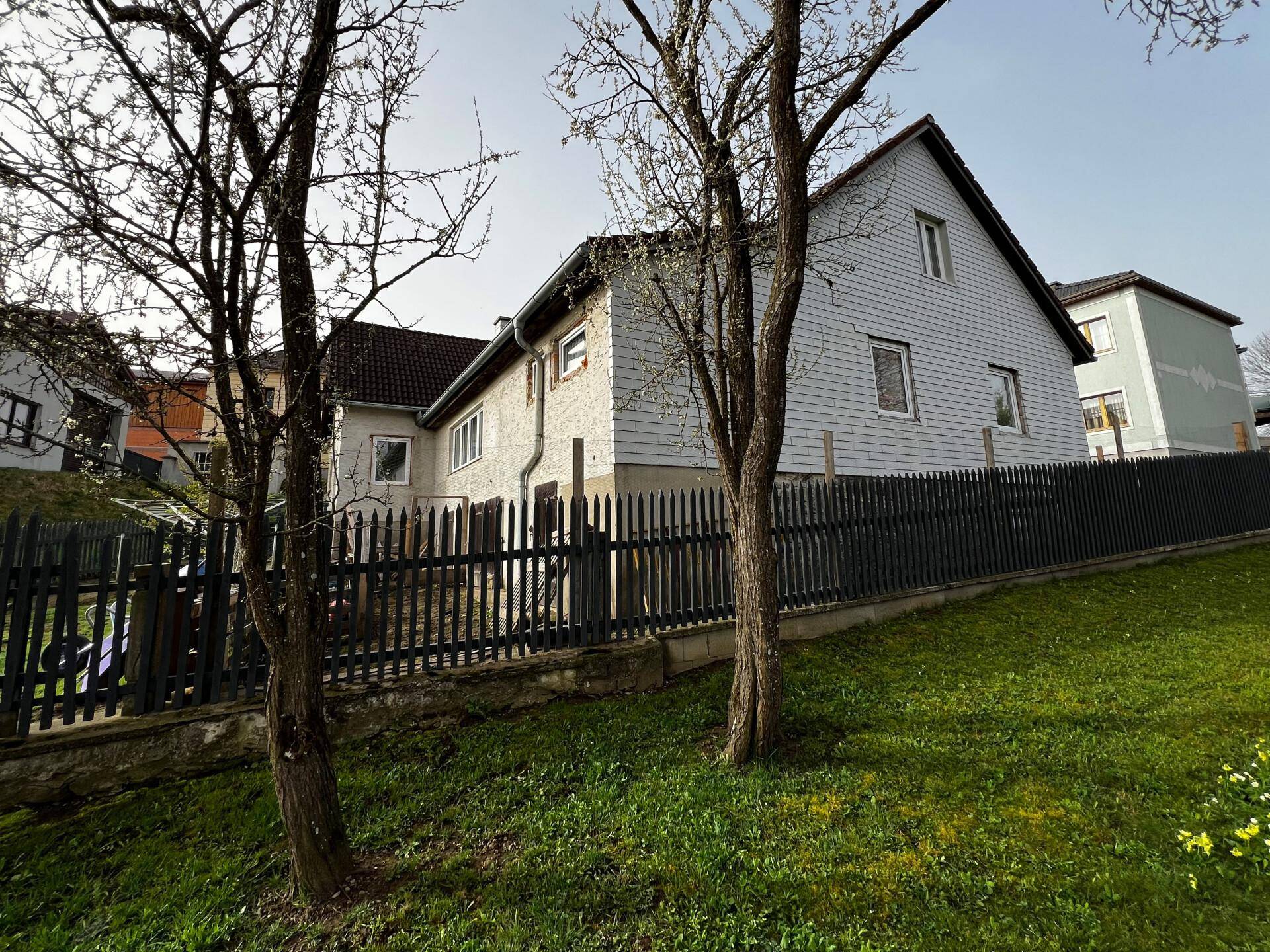 Seitenansicht eines Hauses mit weißer Fassade und dunklem Zaun, umgeben von Bäumen.