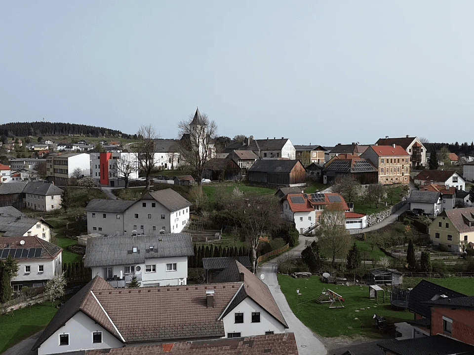 Panoramablick auf ein Dorf mit Häusern, grünen Hügeln und einer Kirche im Hintergrund.